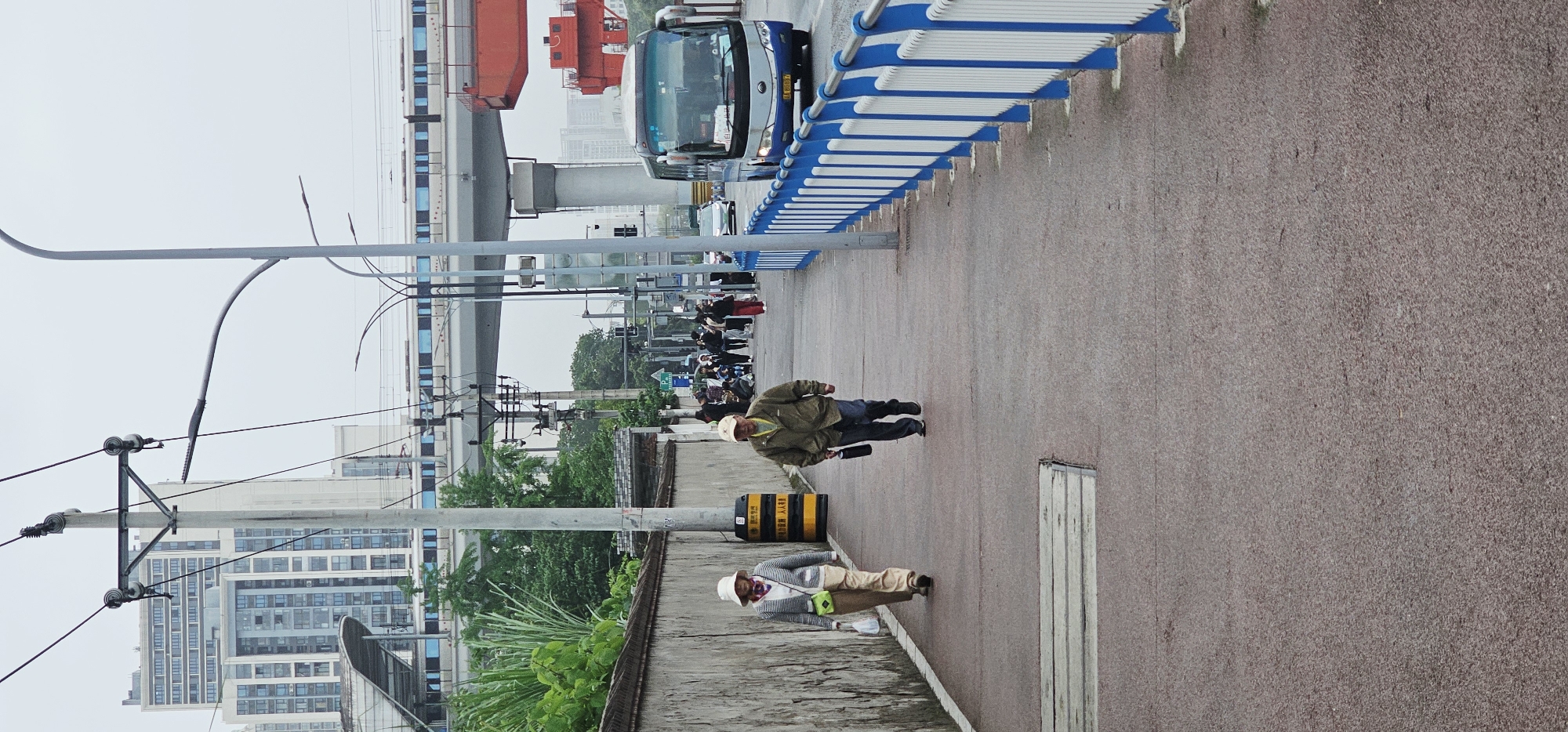 The image depicts an urban street scene in Chongqing High-tech Zone, China, characterized by a paved walkway running alongside a road. An elevated structure, possibly a monorail or highway, spans across the upper part of the frame, parallel to the street level.
In the foreground, two individuals are walking away from the viewer on the paved path. The person closer to the camera wears a khaki jacket, blue jeans, black shoes, and a light-colored hat, carrying a black object in their left hand. The second person, slightly further back, is dressed in a striped shirt, light-colored trousers, a light hat, and carries a bright green bag over their shoulder.
Further down the walkway, a group of people is visible, some appearing to be waiting or congregating. A blue and white public bus is seen driving on the adjacent road, separated from the walkway by a distinctive blue and white striped guardrail. Utility poles with multiple wires line the side of the road and walkway. A black and yellow striped bollard stands on the walkway. In the background, modern multi-story buildings and green foliage are visible under an overcast or hazy sky, suggesting a cloudy daytime.
Visible text includes Chinese characters on the black and yellow bollard that read "施工通道 请勿占用 人人有责," translating to "Construction passage. Please do not occupy. Everyone is responsible." The bus also has a visible license plate, part of which reads "渝D 7***7," indicating its registration in Chongqing.