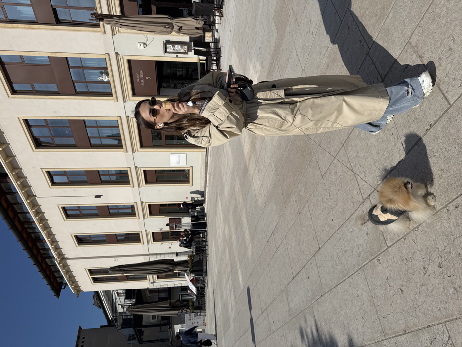 The scene is an outdoor public square in Peschiera del Garda, Italy, in front of the building identified as "Palazzo Alighieri 5". A female subject walks from left to right across a light-colored, patterned flagstone pavement. She has long, dark brown hair, wears small oval sunglasses, and white wireless earbuds. Her attire consists of a long beige trench coat, a plaid shirt visible underneath, light blue ripped jeans, and white chunky sneakers. She is actively eating from a light-colored ice cream cone, held to her mouth with both hands. Her left hand also holds a dark-colored smartphone. A small, light brown Pomeranian-type dog, wearing a black and beige patterned garment, walks on a leash next to and slightly ahead of the woman.
In the background, the multi-story Palazzo Alighieri building features light-colored walls with numerous tall, dark-framed windows and a decorative cornice. The ground floor displays an outdoor dining area with dark tables, chairs, and large, dark umbrella canopies, where several individuals are seated or standing. Further to the left, another person in a blue jacket and white pants walks in the distance. The sky is clear blue, and sharp shadows are cast by the subjects and objects, indicating strong direct sunlight.