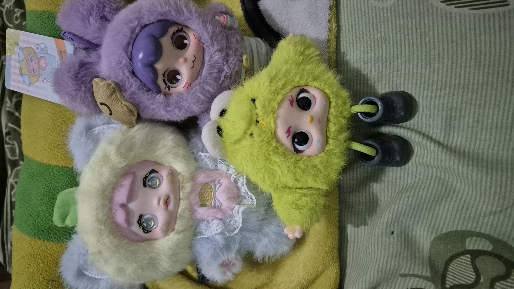 The image shows three plush dolls arranged on a striped fabric surface. The doll on the left has a pale yellow fuzzy hood with grey fuzzy ears, light blue fuzzy body, and a pink face with large blue eyes. The doll in the middle has a purple fuzzy hood with purple fuzzy ears and a brown patch on one ear, a purple face with large brown eyes, and a light grey skirt. The doll in the foreground is lime green and fuzzy, with a pink face, large black eyes, and small black shoes with green laces. The background consists of a yellow and green striped fabric. The scene does not depict any elements that would allow for the determination of its location within the city of Karawang, Indonesia, or any specific actions or interactions.