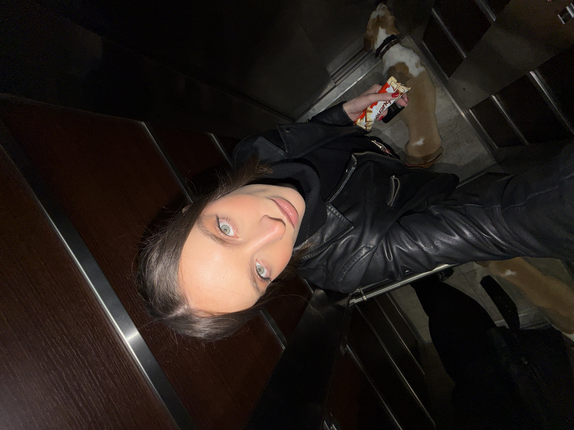 The scene is an interior view of an elevator, captured from a high-angle perspective, within the city context of Budva, Montenegro. A female subject, with brown hair and light eyes, occupies the central frame. She is attired in a black leather jacket over a black turtleneck garment. Her left hand, exhibiting red nail polish, holds a white and red Kinder Bueno chocolate bar. Positioned on the elevator floor to the lower right of the female is a light brown and white dog, wearing a dark collar, lying prone. The elevator's interior walls are constructed of dark wood paneling intersected by horizontal metallic accents. The floor is tiled with light-colored, possibly gray or beige, rectangular tiles. The specific address or precise location within Budva, Montenegro, cannot be identified based on the visual evidence provided in the image.