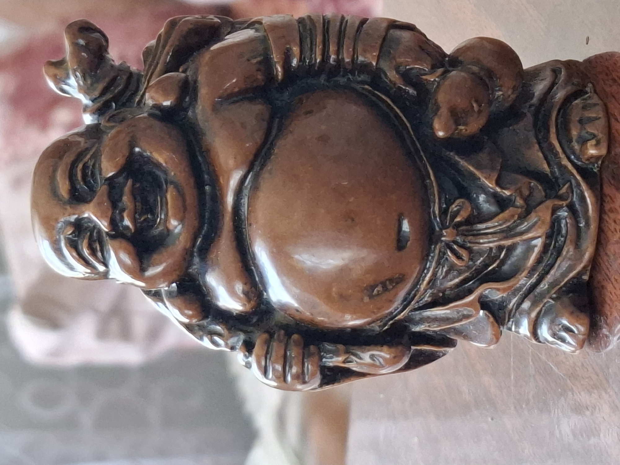 A close-up, eye-level, medium shot shows a small, brown, wooden figurine of a laughing Buddha placed on a wooden surface. The figurine is facing forward and has a large, round belly, a wide smile, and is carrying a sack over its shoulder. The background is out of focus and appears to be a soft, pinkish-brown color. The image does not provide any information about the city of Almaty, Kazakhstan.