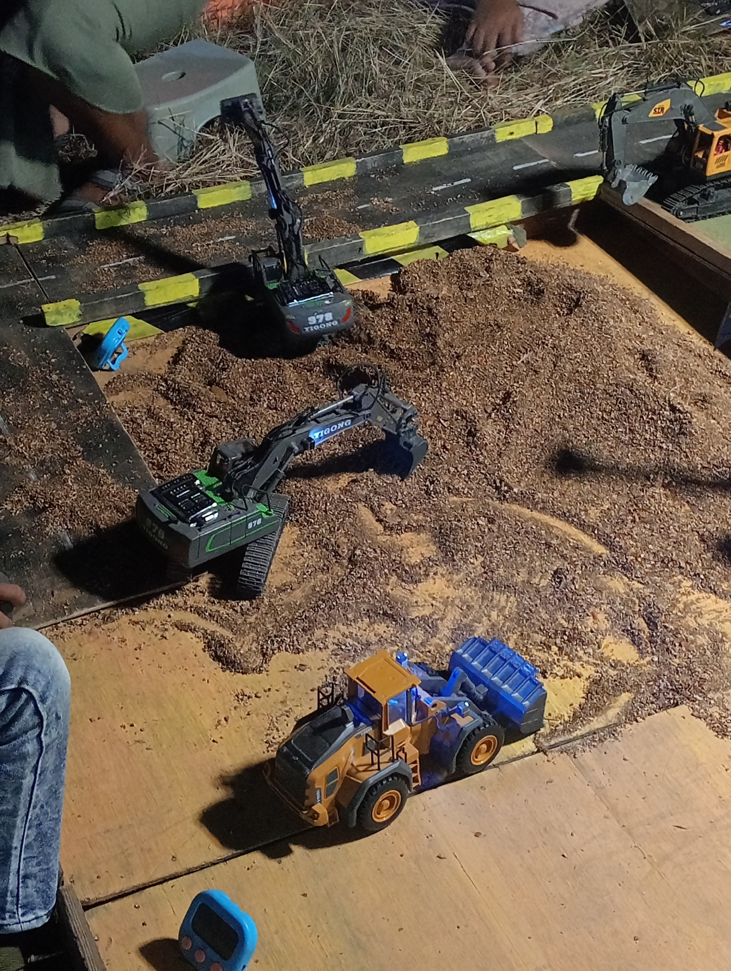 In the village of Ngemplik Wetan, Indonesia, a miniature construction site is set up on a wooden platform. Three toy vehicles are present: a yellow and blue front loader, a black excavator with green accents, and a black excavator with a yellow boom and cab. The ground is covered in a layer of brown, granular material resembling wood chips or dirt. A miniature road, marked with yellow and black safety barriers, runs across the scene. In the background, dry grass and a gray stool are visible, along with a glimpse of a person's legs. The lighting suggests an outdoor setting, possibly at dusk or nighttime.
