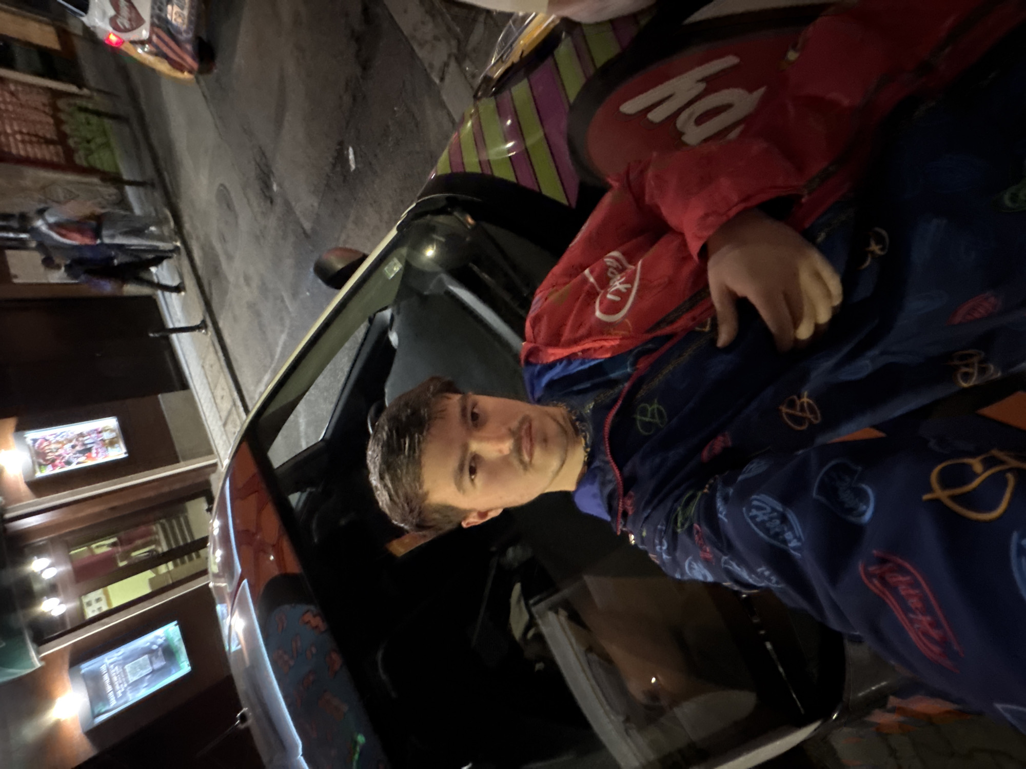 A person with short, dark hair and a mustache sits inside a car. The person wears a blue jacket with printed designs and a red jacket on top. The car's exterior has a multi-colored design. The image is taken in a city environment at night, with street lights illuminating nearby buildings. A building with lit-up signs and displays is visible in the background. The specific location appears to be in Sofia, Bulgaria, based on the design of the car and general cityscape.