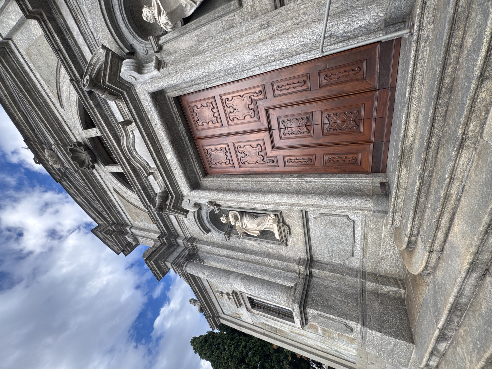 The image depicts the facade of a stone building, possibly a church, with a large wooden double door. The building's exterior is made of rough-hewn gray stone blocks, with intricate carvings and architectural details, including statues in niches. Steps lead up to the entrance. To the left, a tall, dark green evergreen tree is visible, and the sky above is partly cloudy with blue patches. The perspective is from a low angle, looking upwards. This building is located in Balerna, Switzerland.
