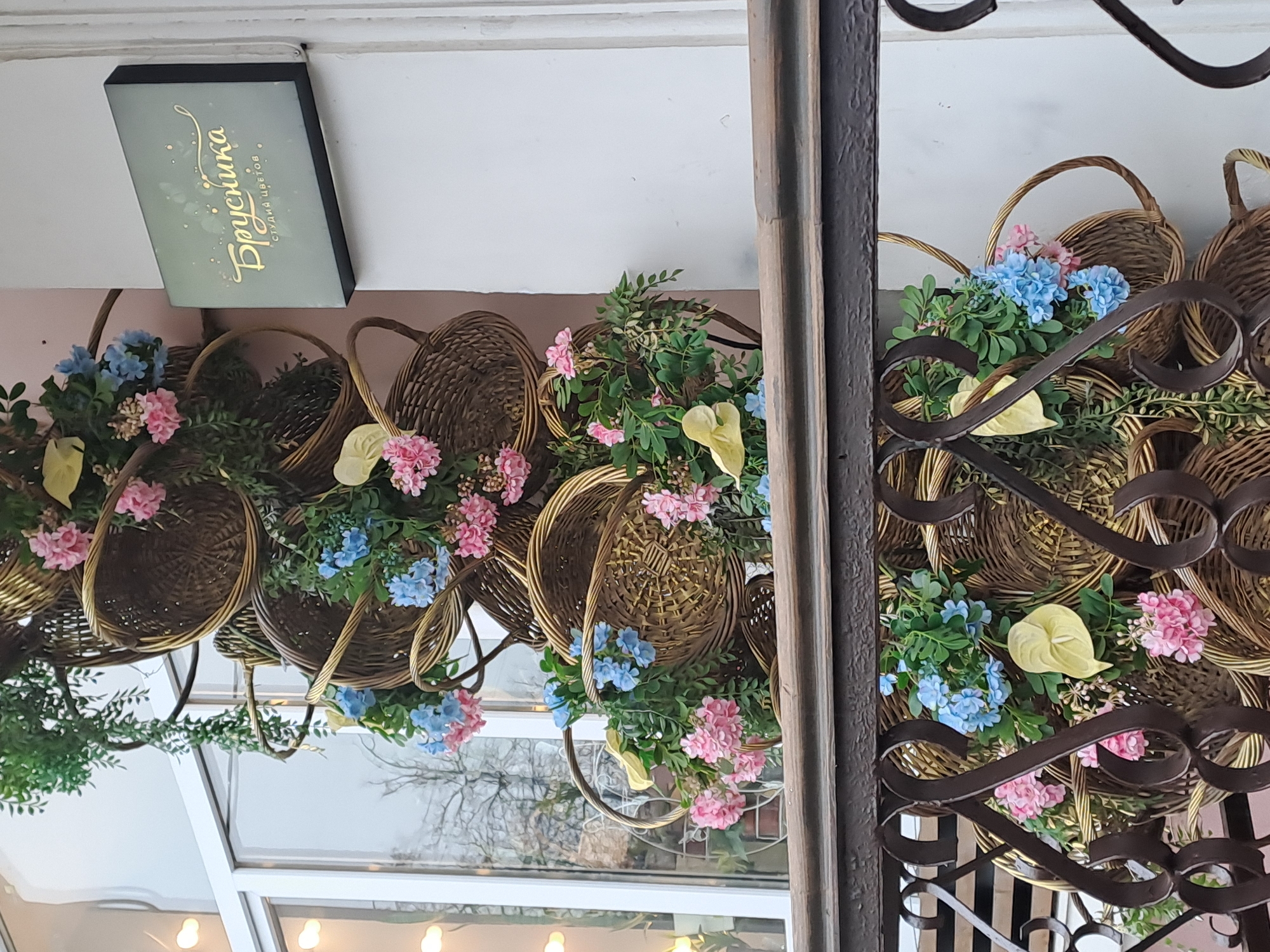 The image shows a shop window or doorway, likely in Elektrostal, Russia, based on the signage. The main decorations consist of several brown wicker baskets filled with colorful flowers, including pink and blue hydrangeas, and light yellow calla lilies. These baskets are arranged along the top of the window and doorway. In the upper left corner, there is a dark rectangle with gold lettering that appears to be a sign for "Брусника Студия цветов" (Brusnika Flower Studio). A decorative metal gate or screen is partially visible on the right side of the frame. The window reflects an outside view, showing trees. The lighting suggests it is daytime.