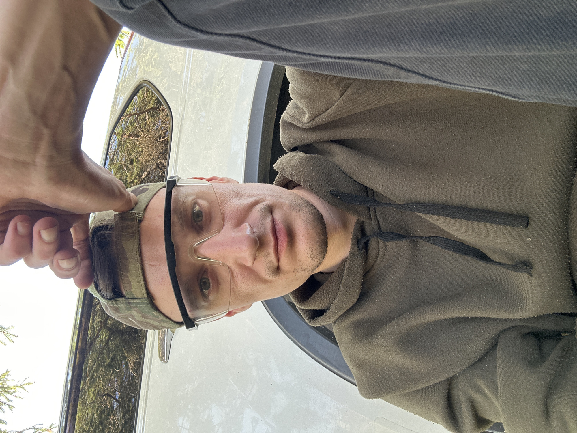 A man wearing a camouflage baseball cap and clear safety glasses is taking a selfie outdoors. He is wearing a green hoodie and a grey long-sleeved shirt. A white vehicle is visible behind him, reflecting trees and the sky. The scene appears to be in a rural or suburban area, possibly near Makiv, Ukraine, given the natural surroundings and the vehicle.