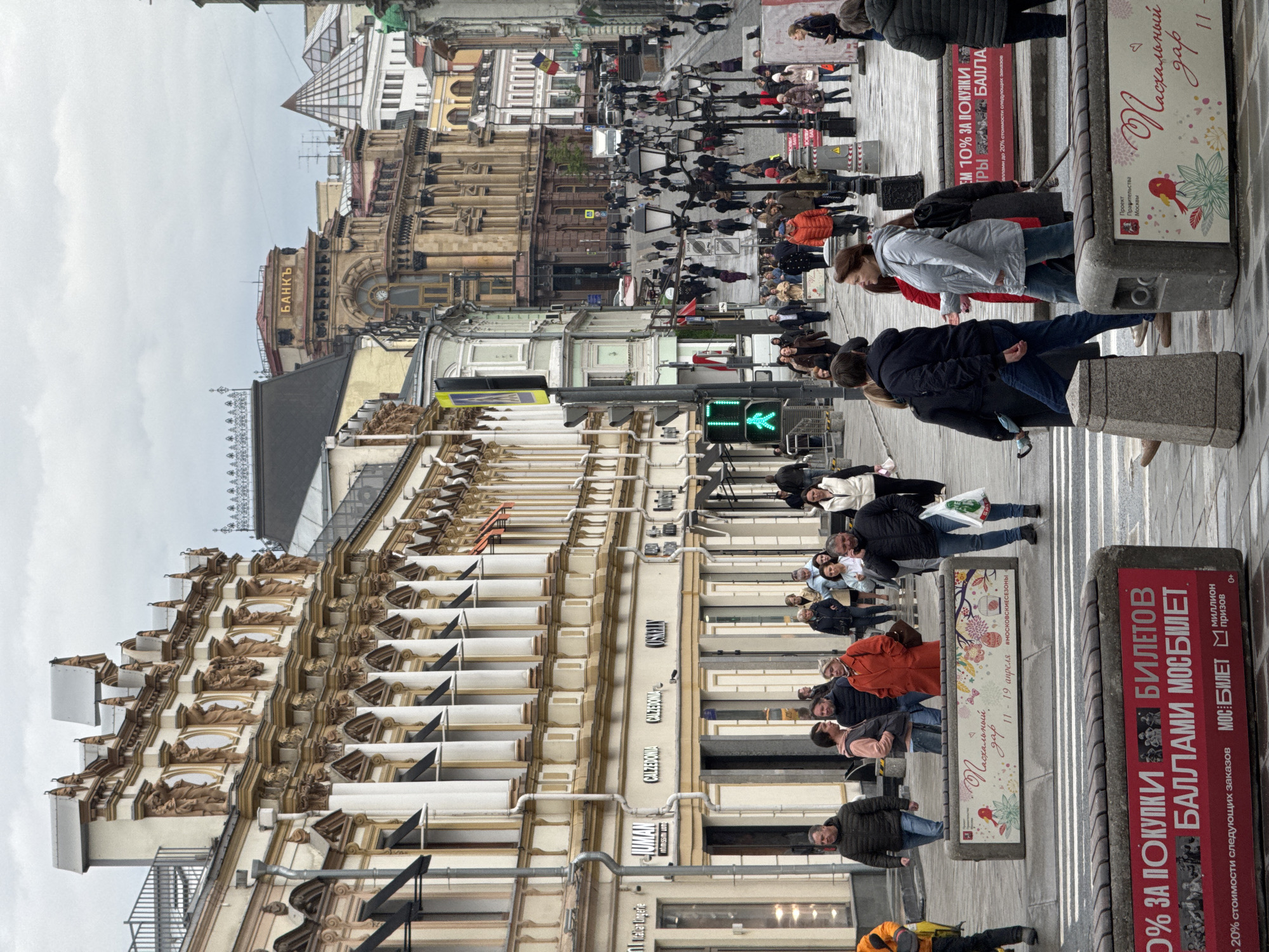 The image captures a bustling pedestrian street in Moscow, Russia, under an overcast daytime sky. Numerous people, dressed in jackets and coats, are seen walking along the sidewalks and crossing a street where a green pedestrian signal is visible.
The street is flanked by ornate, multi-story historical buildings. On the left, a grand building features elaborate architectural details including decorative elements resembling statues. Further down, another building has the old Cyrillic spelling "БАНКЪ" (BANKЪ) displayed prominently. Storefronts are visible, with signs for "CALZEDONIA," "Intimissimi uomo," "JUMAN," "COSKELN," and "Italian Lingerie."
Advertising stands and planters are placed along the sidewalk. A large red sign advertises "30% ЗА ПОКУПКИ БИЛЕТОВ БАЛЛАМИ МОСБИЛЕТ" (30% FOR PURCHASES OF TICKETS WITH MOSBILET POINTS/BONUSES), mentioning "МИЛЛИОН призов" (MILLION prizes) and "до 20% стоимости следующих заказов" (up to 20% of the cost of subsequent orders). Another decorative planter promotes "Пасхальный дар" (Easter Gift) from "11-19 апреля #МОСКОВСКИЕСЕЗОНЫ" (11-19 April #MOSCOWSEASONS), explicitly stating it's a "Проект Правительства Москвы" (Project of the Government of Moscow). The scene depicts everyday urban life in a lively commercial and cultural area of the city.