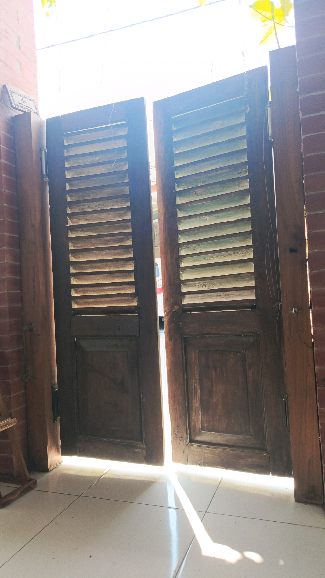 The image depicts an open doorway with wooden louvered doors. Sunlight streams through the gap between the doors and from above, casting a bright light and shadows onto the tiled floor. The doors are made of dark wood, with the upper portions featuring horizontal slats and the lower portions having solid panels. The left door has a hinge visible on its outer edge. To the left of the doorway, a brick wall is visible, with a sign affixed to it. To the right, another brick wall forms the frame of the doorway. The environment appears to be outdoors, likely an entrance to a building or a patio area. The scene is set in Babadan, Indonesia. There are no people visible in the image.