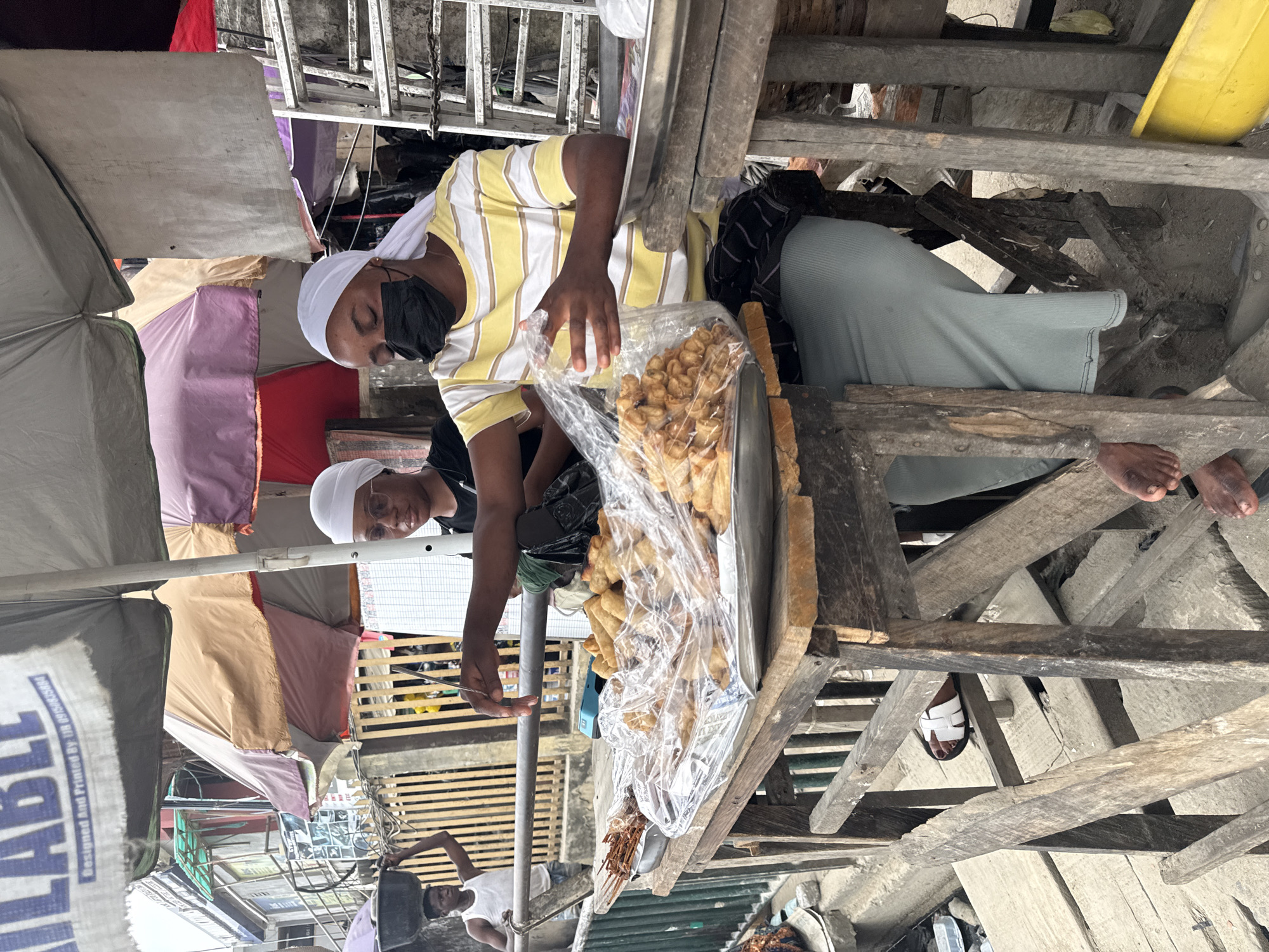 In Lekki, Nigeria, two individuals are seen at a street food stall. One person, wearing a white headscarf and glasses, is interacting with a tray of fried snacks. The other person, also wearing a white headscarf, is behind the stall, wearing a yellow and white striped t-shirt, black face mask, and a light blue skirt. A variety of fried snacks are displayed in plastic packaging on a wooden table. In the background, a man carries a basin on his head, and a ladder is visible against a building. The scene appears to be an outdoor market setting, with umbrellas providing shade.