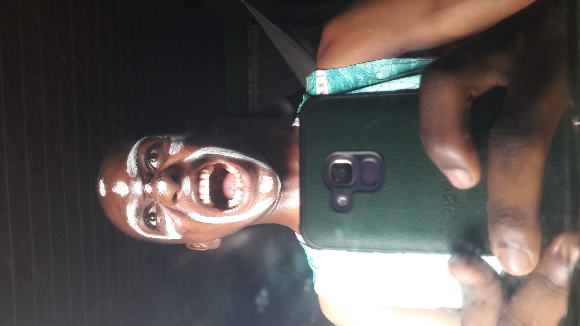 A person with dark skin and white paint markings on their face is holding a dark green smartphone and taking a selfie indoors in Nkpor, Nigeria. The person is looking directly at the camera with their mouth open, revealing their teeth and tongue. The phone's camera and flash are visible. A portion of the person's shoulder and blue and green patterned clothing are visible, as well as a window with white curtains. In the background, dark brown striped walls can be seen.