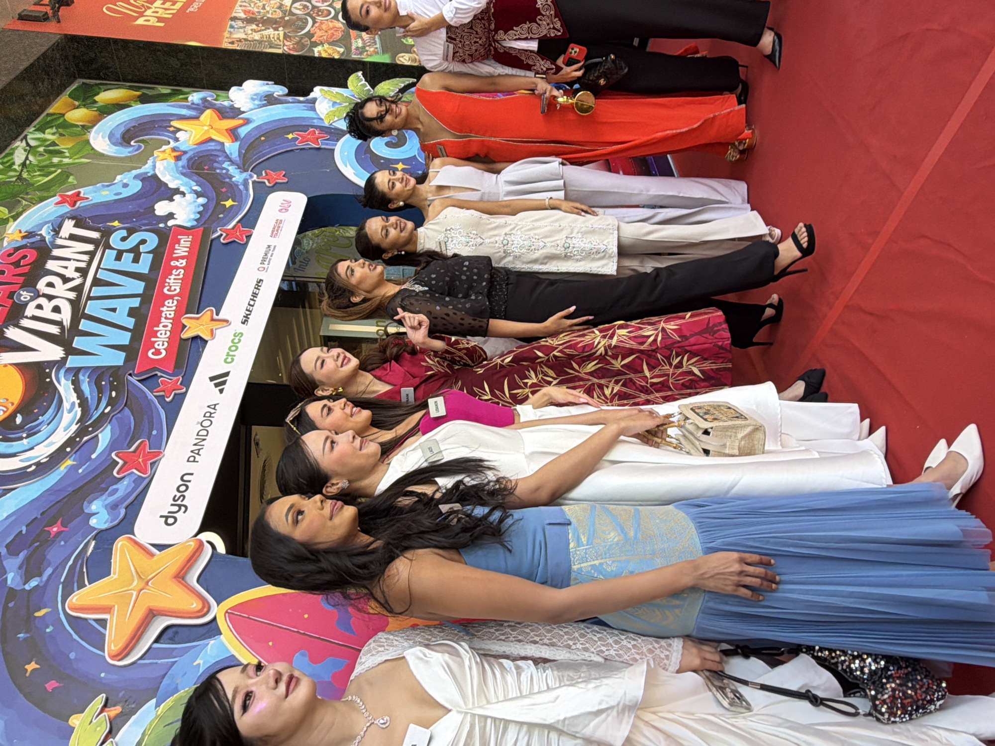 The photograph captures a group of eleven women standing on a red carpet in front of a vibrant backdrop. The backdrop features a large banner with the text "VIBRANT WAVES" prominently displayed, alongside imagery of stylized waves, stars, and palm trees. Several sponsor logos, including "Dyson," "Pandora," and "Adidas," are visible on the backdrop. The women are dressed in a variety of formal and semi-formal attire, ranging from long gowns to elegant pantsuits. They appear to be participating in an event or competition, as some have name tags pinned to their clothing. The overall setting suggests an outdoor event or an open area within a building, possibly a mall or convention center, given the large banner and polished flooring. The lighting suggests it is daytime, with some artificial lighting also present. The women are posing for the camera, with some smiling and others looking towards the lens. The event appears to be festive and celebratory. The context of Nha Trang, Vietnam, implies that this event might be related to a local festival, tourism promotion, or a beauty pageant, given the demographic of the participants and the celebratory atmosphere.