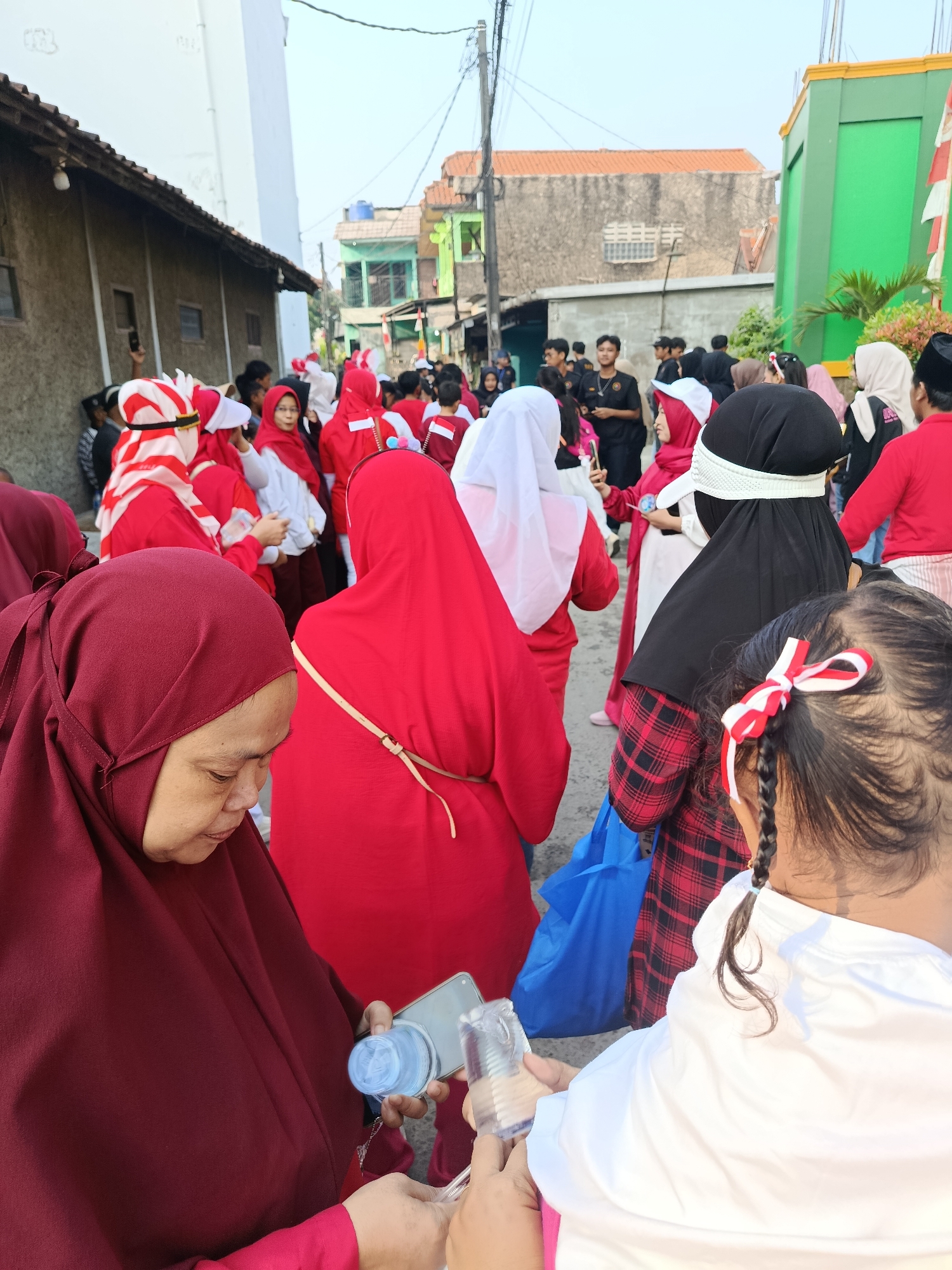 In the city of Balaraja, Indonesia, a group of people, predominantly women wearing red and white attire, are gathered outdoors, possibly for a local celebration or event. The scene appears to be a narrow street, with buildings lining both sides. The people are standing close together, some are holding phones, and one woman in the foreground is interacting with a bottle of water and her phone. The attire suggests a patriotic theme, with red and white being the national colors of Indonesia. The overall atmosphere seems festive and communal.