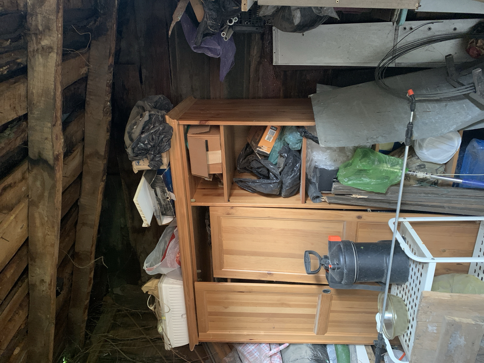 The image shows a cluttered storage space, likely inside a shed or attic, in an unknown location. The space features rough, unfinished wooden planks forming the walls and ceiling. A wooden dresser with drawers is visible, filled with boxes, garbage bags, and other miscellaneous items. Additional objects, including metal sheets, a white plastic crate, a black water tank, and a spray bottle, are piled around and on top of the dresser. The overall environment is disorganized and dimly lit.
