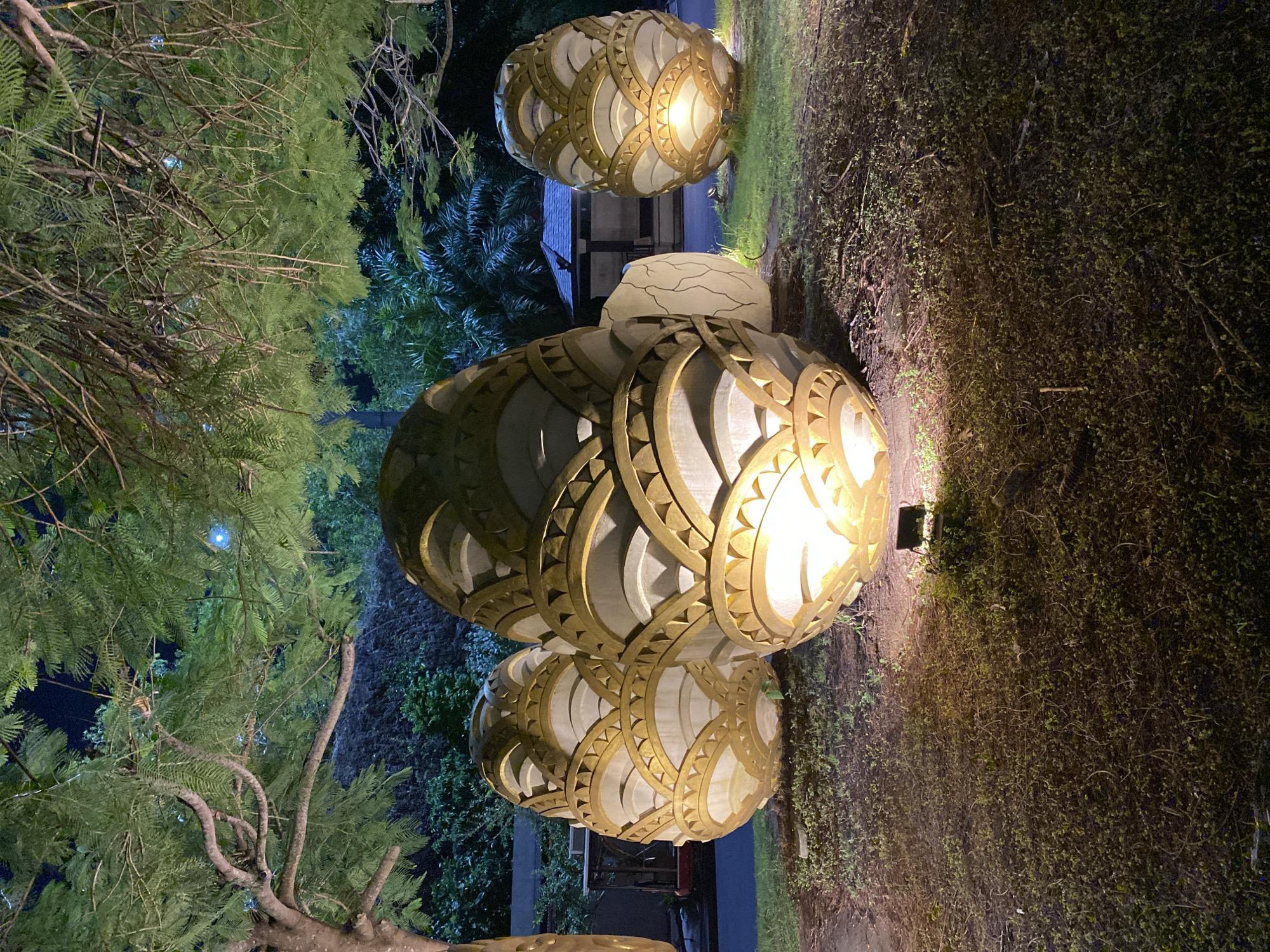 In Kuta Selatan, Indonesia, at night, four large, egg-shaped sculptures are illuminated by spotlights from the ground. The sculptures are made of a white material with gold, fan-like decorative elements arranged in concentric layers. Three sculptures are clustered together in the foreground, with a fourth sculpture visible to the right, slightly further back. The largest sculpture is in the center foreground. Behind the sculptures, a small, dark-roofed building with white walls and dark trim is visible. Lush green trees with dense foliage dominate the upper portion of the image, their branches casting shadows and framing the scene. The ground is covered in grass and dry leaves. The overall atmosphere is serene and illuminated, suggesting an outdoor art installation.