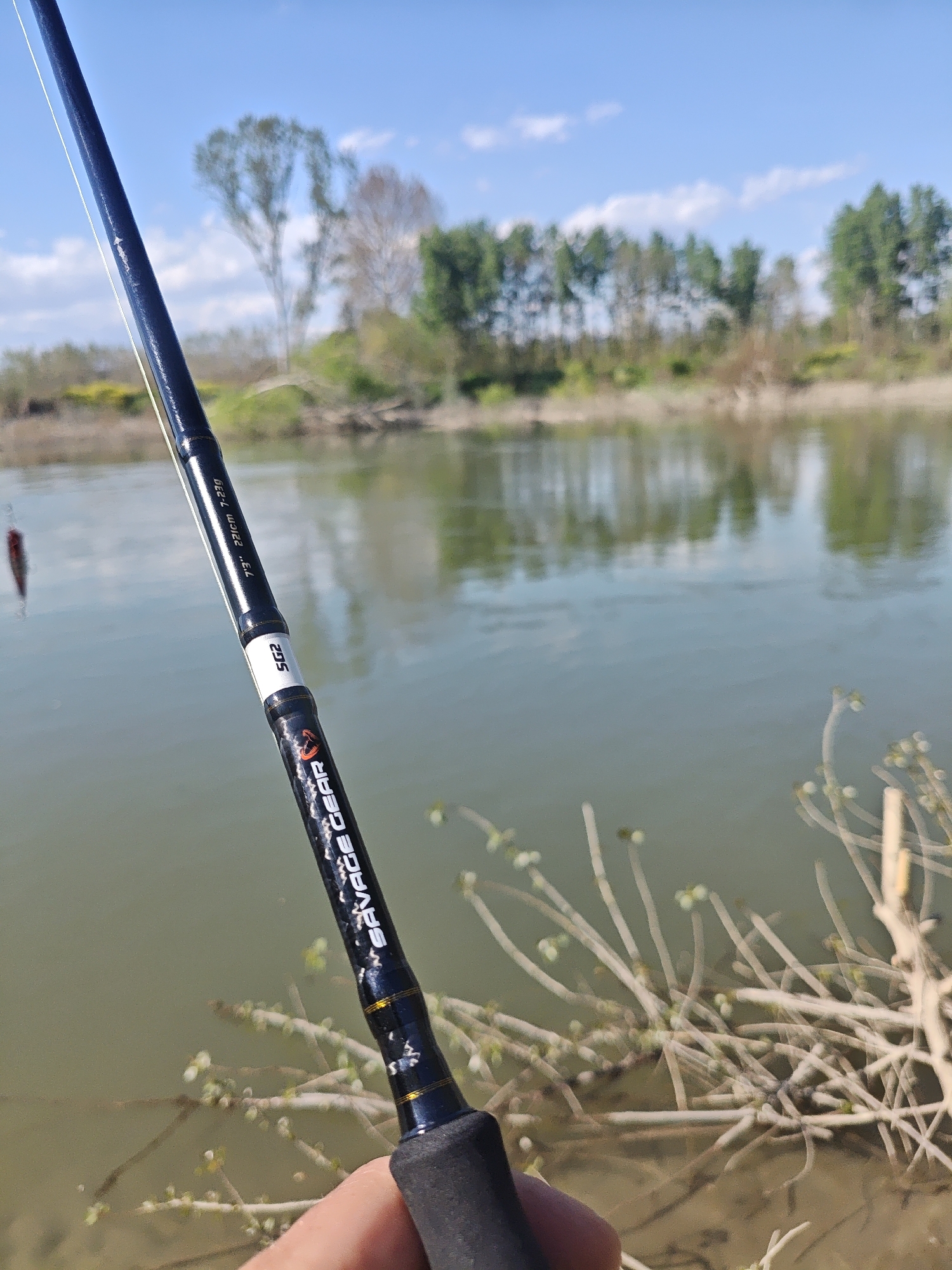 A person is holding a black Savage Gear fishing rod, 7'3" 221cm 7-23g, with SG2 branding, near a river. The river reflects trees on the opposite bank. Dry brush is visible near the riverbank. The sky is blue with scattered clouds. This scene is located in the Municipal Unit of Skoutari, Greece.
