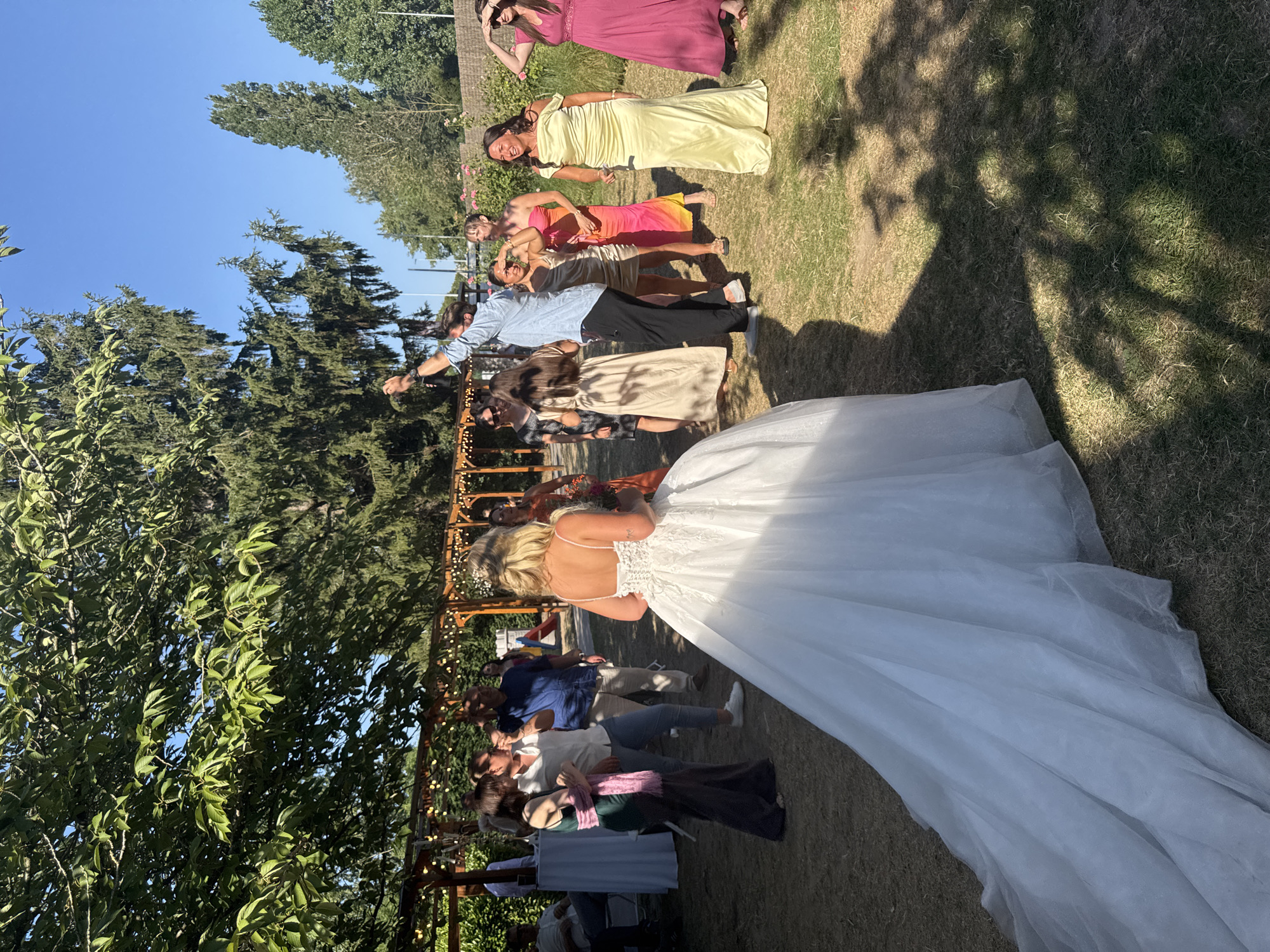 In Hem, France, a wedding reception is underway outdoors under a clear blue sky. A bride in a white gown stands with her back to the camera, her train spread across the lawn. Guests mingle in the background, some in formal attire, others more casually dressed. A man in a light blue shirt and dark pants is seen with his arm raised. The scene is set in a grassy area with trees and a wooden pergola adorned with string lights.
