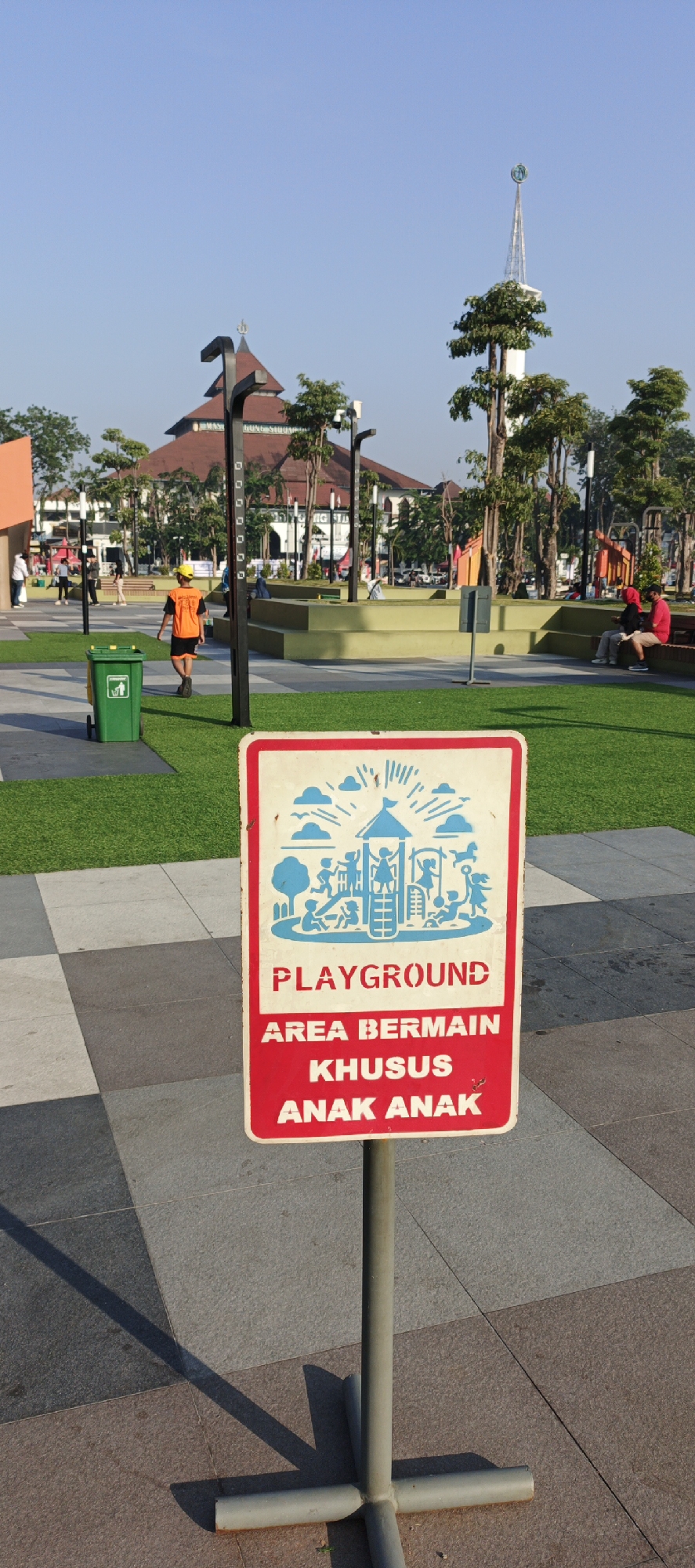 In Sidoarjo, Indonesia, a clear sky presides over a public outdoor space. A prominent sign, red with a white border, stands on a metal pole in the foreground, its text in red and blue. The sign features a stylized illustration of a playground with children playing, and reads in English: "PLAYGROUND AREA BERMAIN KHUSUS ANAK ANAK". Behind the sign, a paved area transitions to a manicured lawn. A green trash can with a recycling symbol is visible on the lawn to the left. In the middle ground, a person in an orange vest and yellow hard hat walks away from the camera towards the left. Beyond this, a large building with a distinctive tiered roof, possibly a mosque given its architecture, is visible, adorned with a green and white spire. Several trees with lush green foliage are scattered throughout the scene, interspersed with streetlights and what appear to be low-rise seating structures. In the background, more trees and distant buildings can be seen, along with a few individuals seated and walking, indicating a leisurely public atmosphere. The overall impression is one of a well-maintained recreational area under a bright, sunny sky.