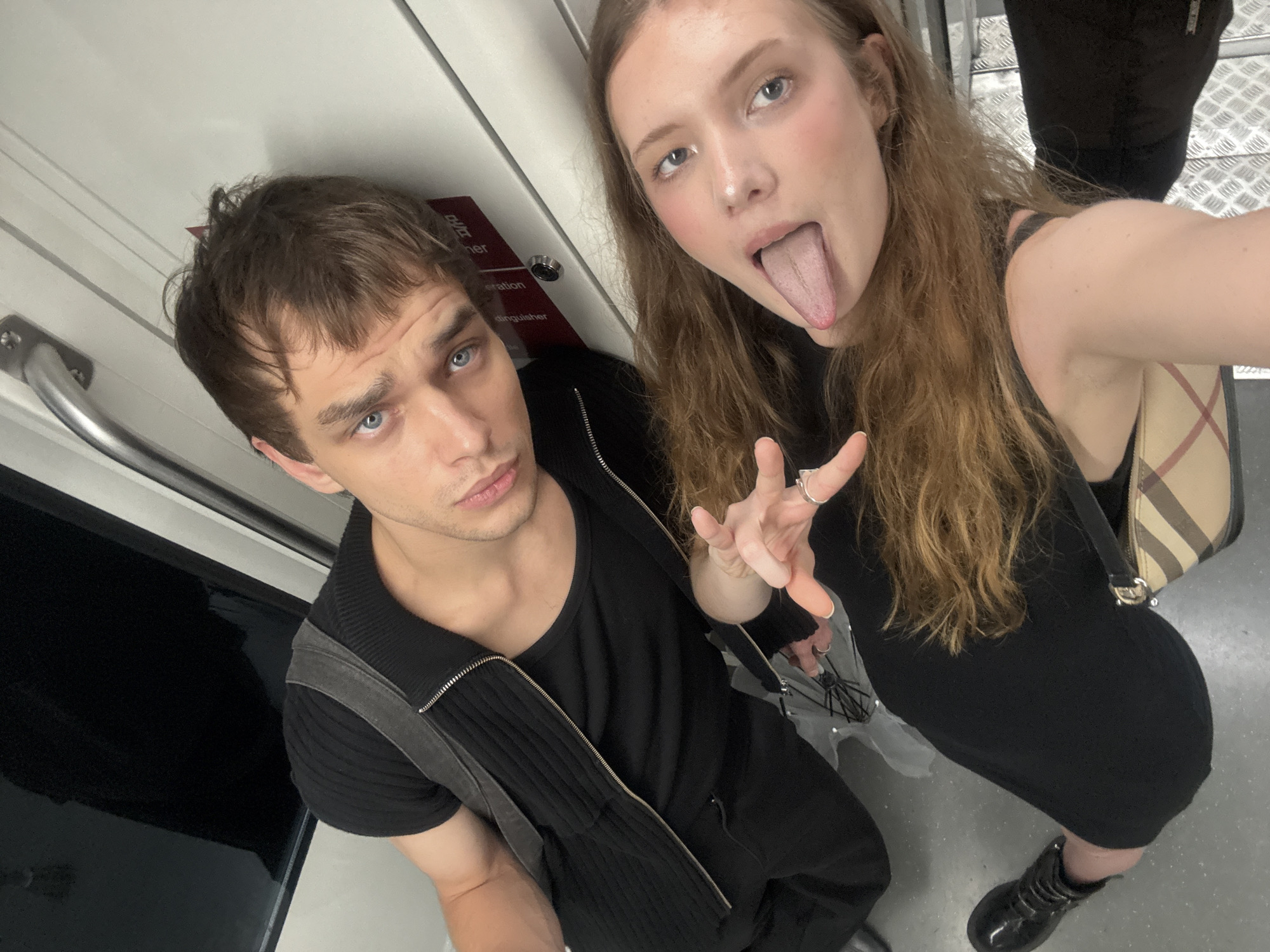 In the city of Xuhui District, China, two individuals, a man and a woman, are captured in a close-up selfie taken from a high angle, likely inside a mode of public transportation, suggested by the metallic handrail and patterned flooring visible. The man, positioned on the left, has short brown hair and striking blue eyes. He is wearing a black t-shirt layered with a black zipped vest. The woman, to the right, has long, light brown hair and is sticking her tongue out towards the camera, with a ring on her left index finger. She is wearing a black dress and a distinctive checkered shoulder bag. The background appears to be the interior of a train or subway car, indicated by the door panel with red signage and a metal handrail.