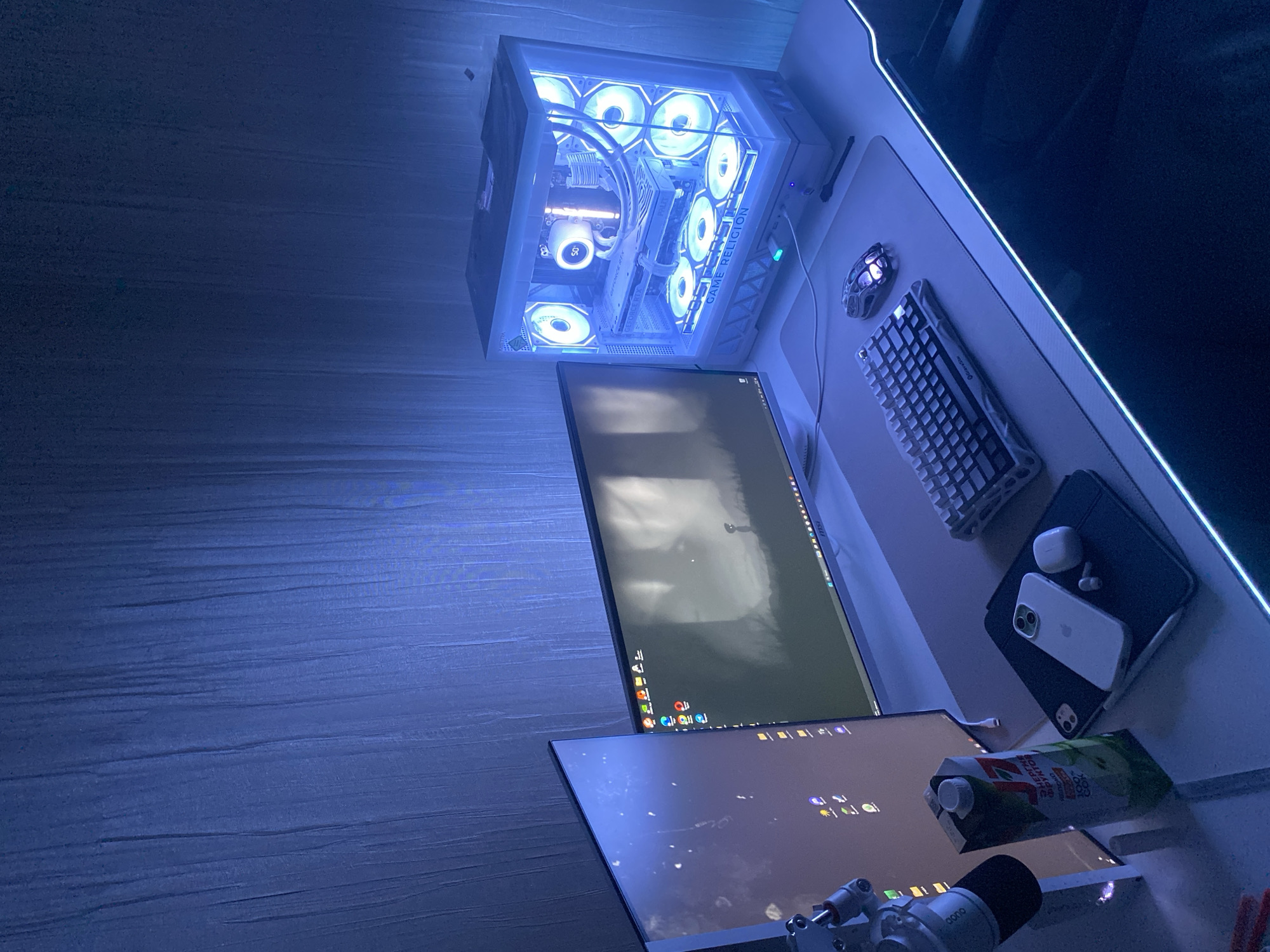 This image captures a brightly lit, modern gaming or computer setup on a desk, likely in a room in Chaikovsky, Russia, during nighttime or in a dim environment, as indicated by the glowing components. The dominant color scheme is cool white and blue, emanating from the PC and desk lighting.
The main subject is a high-performance desktop computer tower, white with a transparent side panel, showcasing numerous LED-lit fans, water cooling, and a white "GEFORCE RTX" graphics card. A display on the CPU cooler shows "56". Prominently visible on the PC case is the illuminated text "GAME RELIGION".
The setup includes two monitors, with desktop icons visible on both screens, suggesting active use. A white mechanical keyboard, faintly displaying "CHERRY", rests on a large, light-colored desk mat next to a patterned gaming mouse. Other personal electronics on the desk include a white iPhone with a light green camera module, a pair of white AirPods in their case, and a dark-colored tablet, possibly an iPad.
A carton of beverage, partially showing Cyrillic text "ЗЕЛЁНЫЙ... ...СОК" (Green... ...Juice) and "100% сок" (100% juice), is placed near the tablet. A white microphone on a boom arm is visible in the foreground. The overall scene suggests a dedicated and well-equipped personal computer workstation for gaming or productivity.