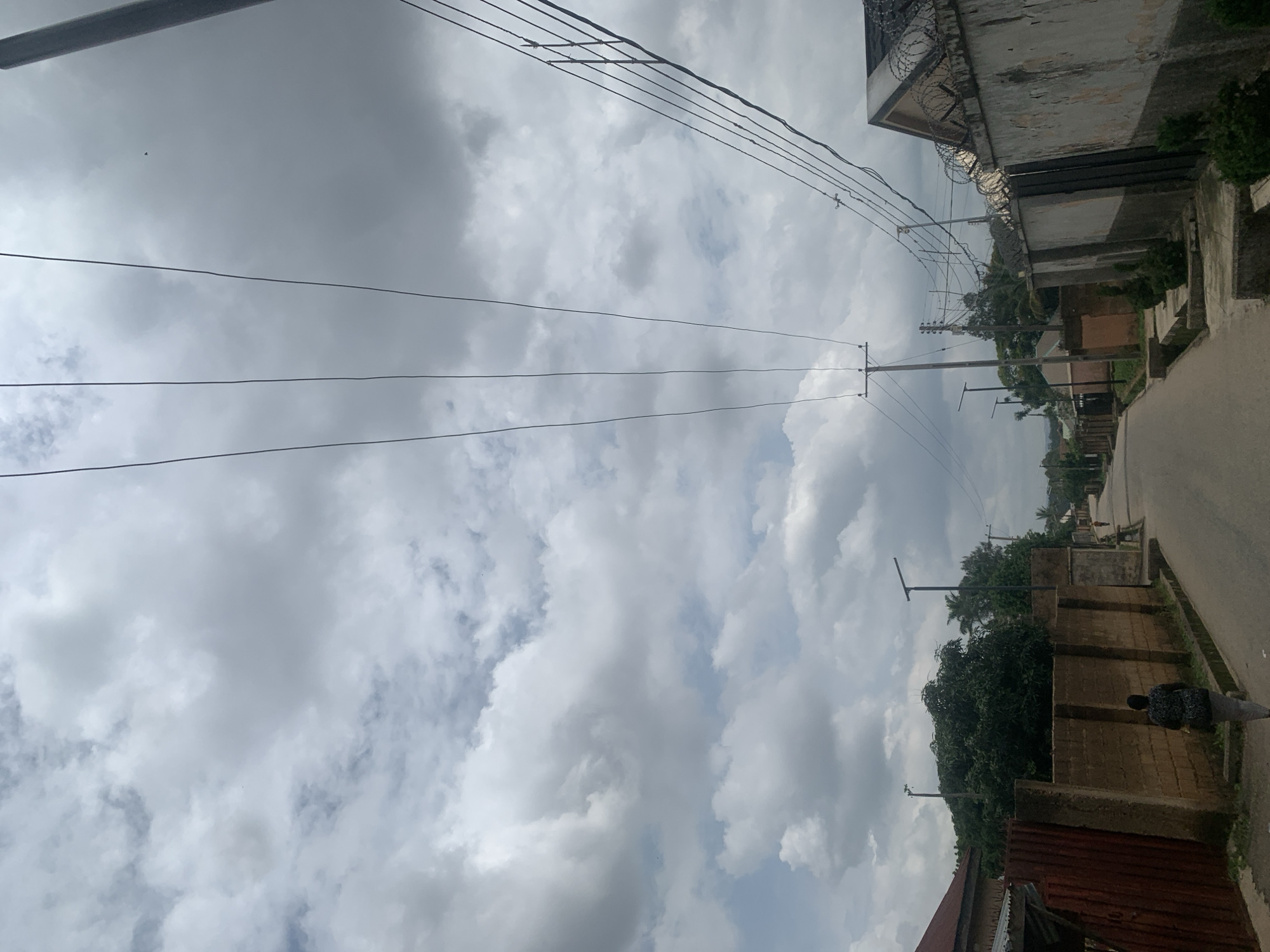 This is a vertical, eye-level, full-shot photo of a street scene in Jos, Nigeria. The sky is filled with dense, grey clouds, suggesting an overcast or potentially rainy day. Multiple power lines stretch across the frame, intersecting the sky and extending towards utility poles visible in the distance.
The scene depicts a street running alongside buildings and a wall. The buildings appear to be made of concrete and brick, with some showing signs of wear and tear. One building on the right has barbed wire along its top, hinting at security measures. Vegetation, including trees and bushes, is present, adding a touch of green to the otherwise muted color palette.
A person, wearing a patterned shirt and grey pants, is walking away from the camera on the sidewalk. They are the only clearly visible person in the frame. The road surface is asphalt. The overall impression is one of a typical day in a residential or semi-urban area, possibly with a slightly somber atmosphere due to the cloudy sky. There is no visible text in the image. The time of day appears to be daytime, but the heavy cloud cover obscures any clear indication of sunlight.
