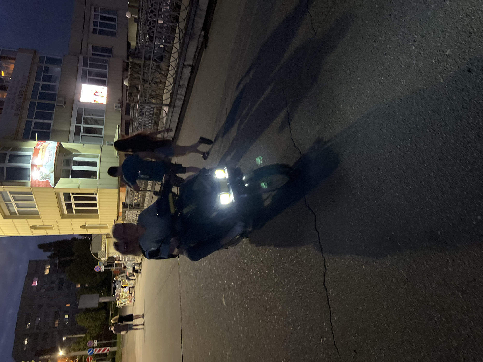 In Krasnodar, Russia, at dusk, a couple rides a scooter on a cracked asphalt road. The scooter's headlights illuminate the path ahead. In the background, a multi-story building with illuminated windows stands tall, and a fence with decorative metalwork lines the side of the road. Several other individuals are visible walking in the distance. The overall scene captures a moment of evening activity in an urban environment.