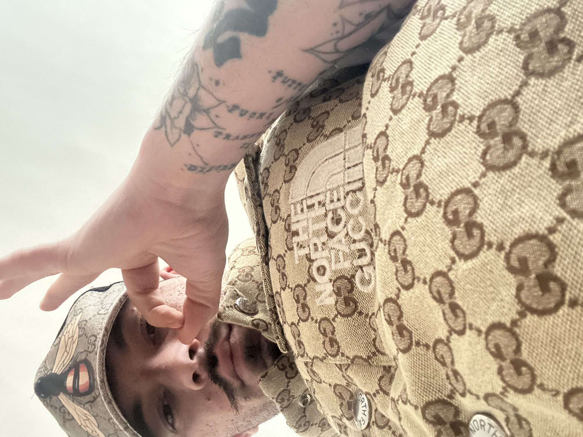 A male individual is depicted from a low-angle perspective, looking directly down at the camera. He possesses a dark, styled handlebar mustache. His left hand is raised to face level, forming an "OK" gesture with the thumb and forefinger, positioned to the right of his nose. He wears a beige baseball cap adorned with a prominent embroidered bee design over a repeating "GG" pattern. His outer garment, likely a jacket or vest, is made of beige fabric patterned with repeating brown "GG" logos. On the chest, embroidered in white, are the words "THE NORTH FACE GUCCI." Metal snap buttons on the garment feature "THE NORTH FACE" text. The subject's right forearm is visible, extensively covered with black and grey tattoos including floral designs and script. The background is a plain, undifferentiated light surface, providing no specific environmental details. The exact location within the city of Varna, Bulgaria, cannot be determined from the visual information provided.