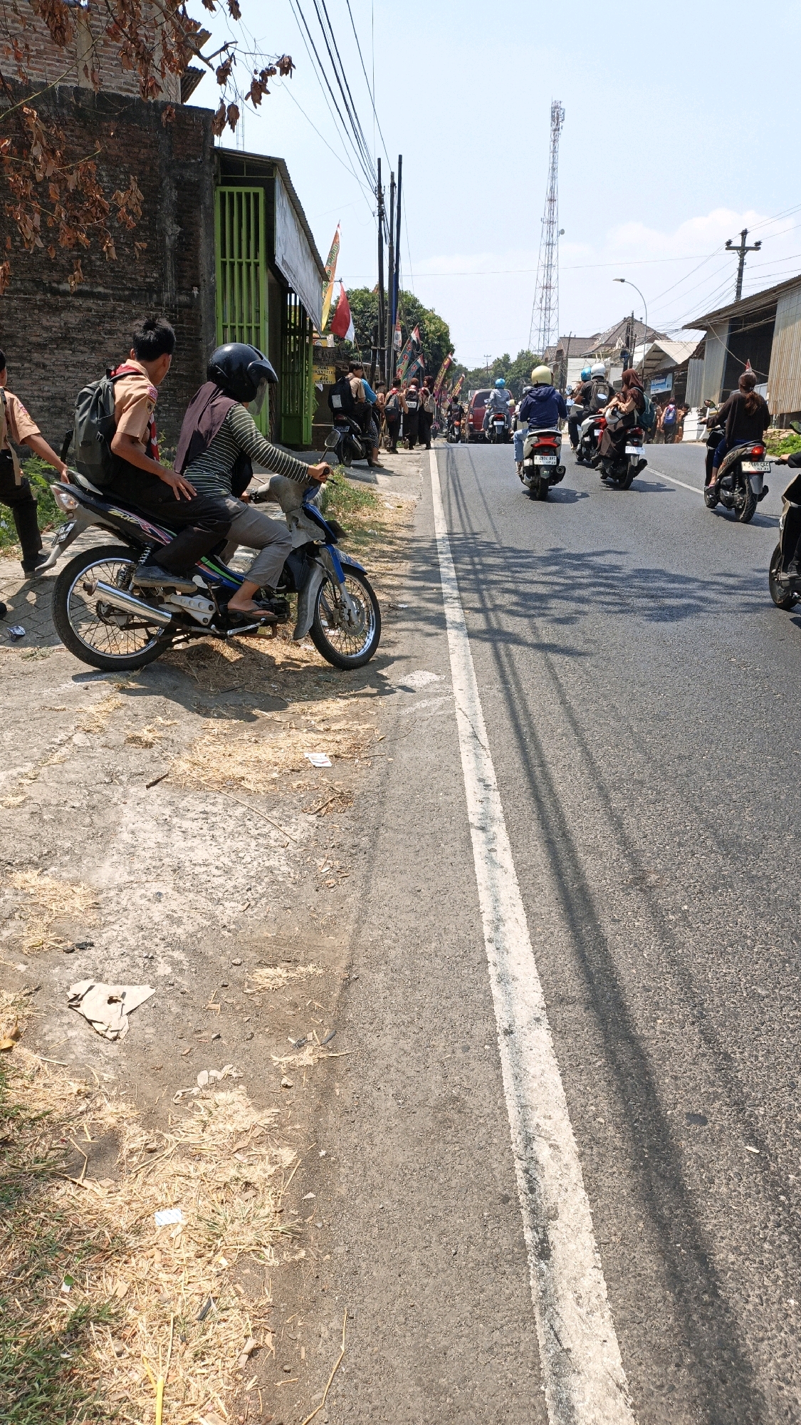 In Kuwasen, Indonesia, a street scene unfolds on a sunny day with a clear sky. The road surface is asphalt, marked by a white line dividing the lanes, and shaded by intermittent tree shadows. To the left, a dirt and grass embankment lines the road. A brick building with green barred doors and windows stands on the embankment, adorned with Indonesian flags. Adjacent to it are more buildings and a cellular tower in the distance.
Several people are present, mostly on motorcycles. In the foreground, two individuals, possibly students in scout uniforms, are on a blue motorcycle parked on the side of the road. The rider wears a helmet and a striped shirt, with the passenger in the back carrying a backpack. Further down the road, a procession of motorcycles is visible, carrying various riders and passengers, some with backpacks. The overall atmosphere suggests a gathering or a journey.