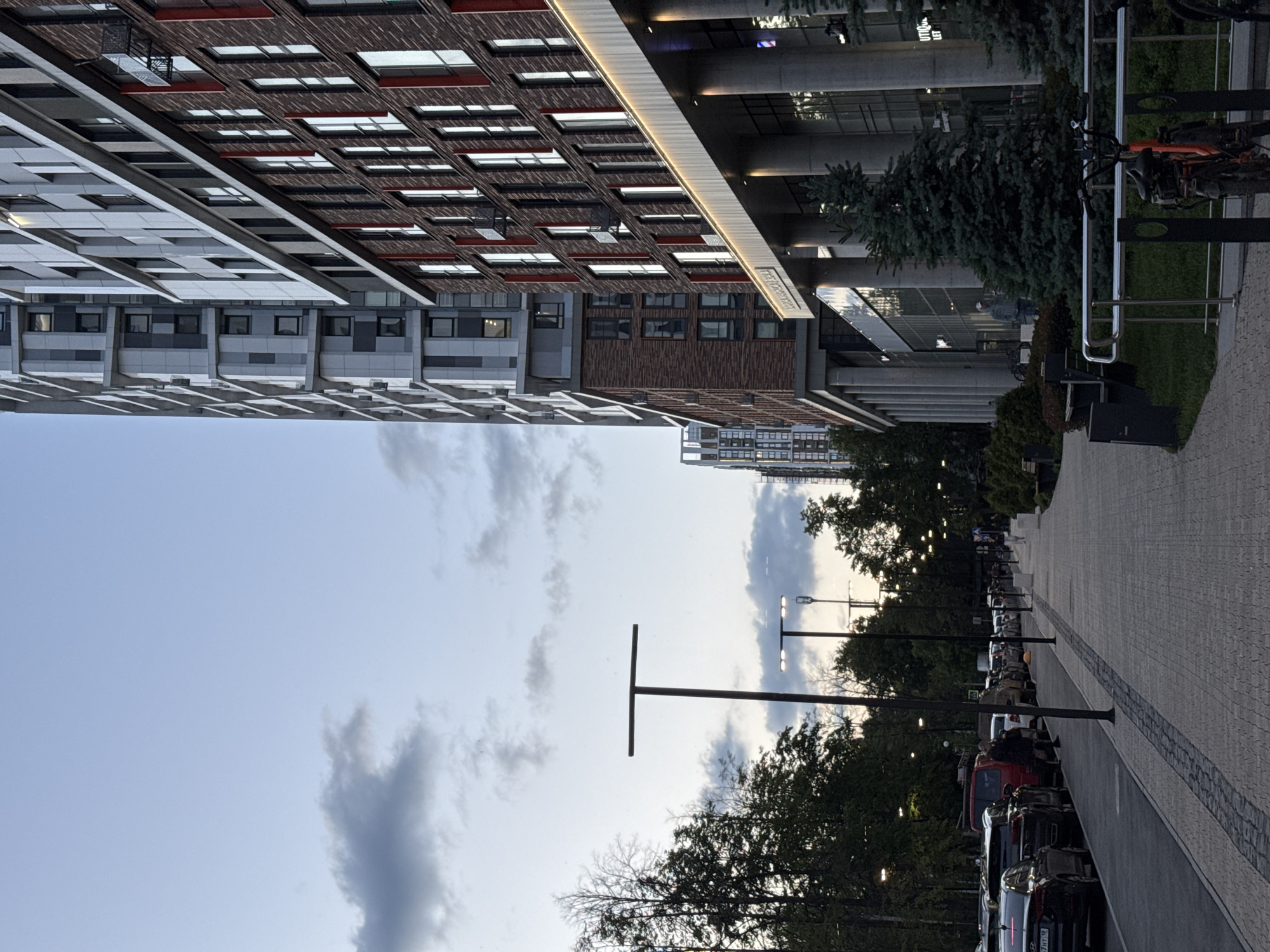 The image captures a modern urban street scene, likely in the late afternoon or early evening, characterized by soft natural light and illuminated architectural features. The setting includes contemporary buildings, a paved sidewalk, a street lined with parked cars, and various trees, all under a partially cloudy sky.
On the left, several multi-story buildings dominate the view. One prominent structure features an intricate facade with reddish-brown brick-like patterns interspersed with numerous rectangular windows. Adjacent to it, another building displays a pattern of light grey and white panels, also with many windows. A white, illuminated ledge runs along one of the buildings, casting a subtle glow.
The right side of the frame shows a wide, paved sidewalk made of grey interlocking pavers, bordered by a strip of grass and various trees, including an evergreen conifer. Beyond the sidewalk, a street is filled with numerous parked cars, including a red van and several dark-colored vehicles. Modern streetlights are positioned along the road, though not all appear to be fully lit. A bike rack with bicycles is visible near one of the building entrances.
The sky above is light blue with scattered grey clouds, suggesting either dusk or an overcast day. A few blurry figures can be discerned walking near the building entrances and on the sidewalk, indicating typical pedestrian activity. No prominent text is clearly legible, though faint stylized letters can be observed on the illuminated building ledge and possibly above an entrance. The overall atmosphere is calm, depicting a typical urban environment in Izhevsk, Russia.