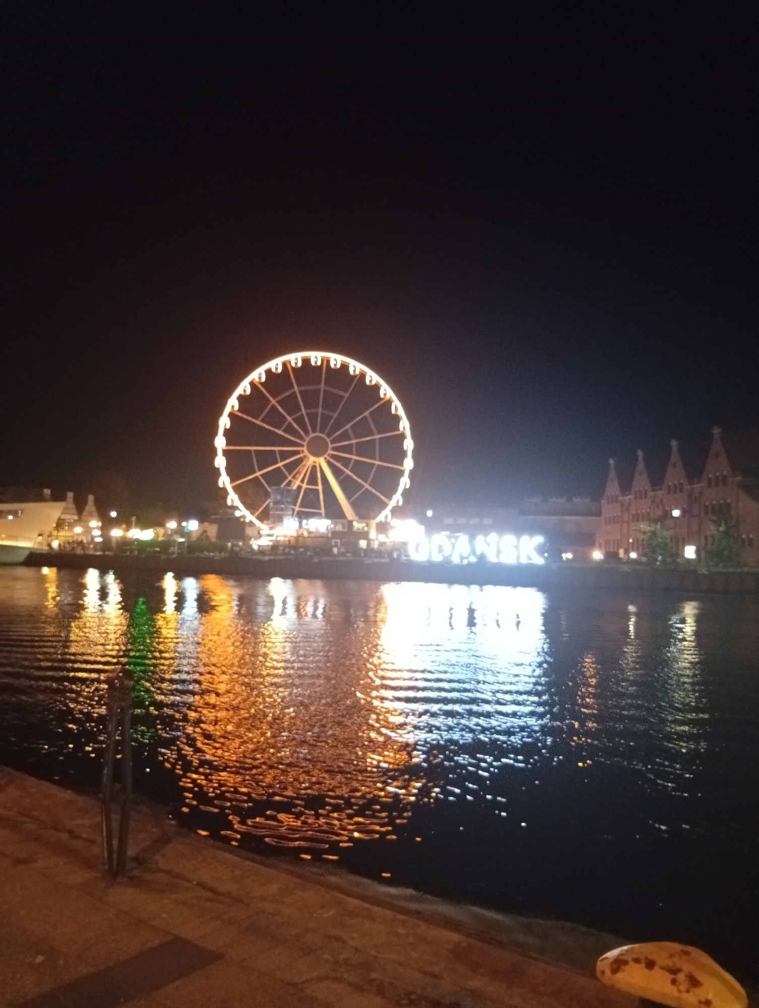 The image captures a nighttime view of the Motława River in Gdańsk, Poland. A large Ferris wheel illuminated with white lights stands prominently on the waterfront, reflecting in the dark water. To the right of the Ferris wheel, large white illuminated letters spell out "GDAŃSK," serving as a landmark. Behind the letters, a historic brick building with arched windows and a distinct gable roofline is visible. The river's surface is rippled, mirroring the lights from the Ferris wheel and the city. In the foreground, the edge of a wooden pier or embankment is visible, with a single green illuminated object reflecting in the water, possibly a small tree or decoration. The overall scene is a lively waterfront at night, showcasing iconic elements of the city.