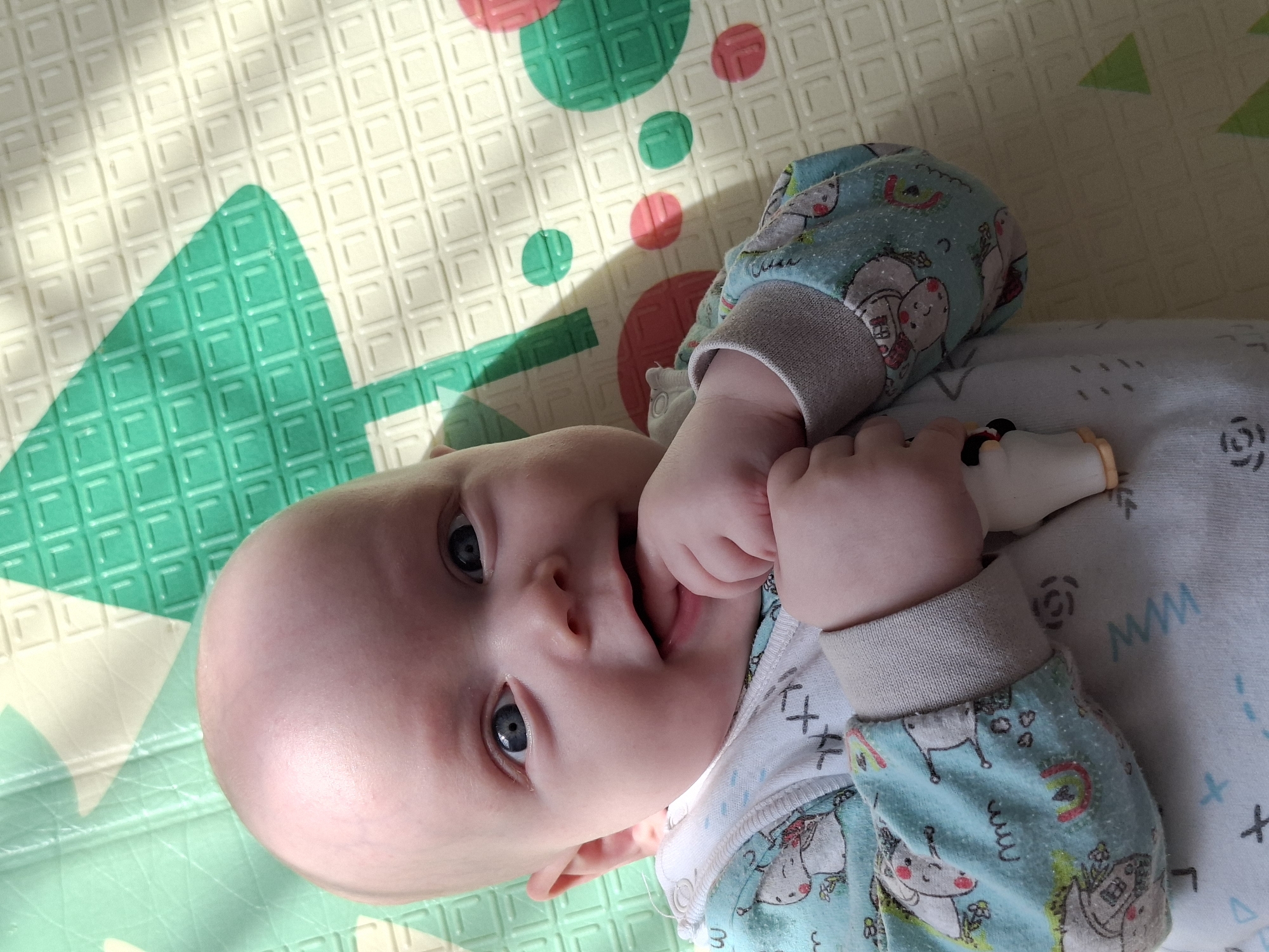 A baby lies on a padded play mat with a green and pink geometric pattern. The baby is wearing a light blue and white patterned onesie with gray cuffs. The baby's eyes are open and focused towards the viewer. The baby has a light-colored complexion and a bald head. The baby's hand is near their mouth, and the fingers are partially inside the mouth. The baby is holding a small, light-colored toy. The setting is indoors. The location is unknown.