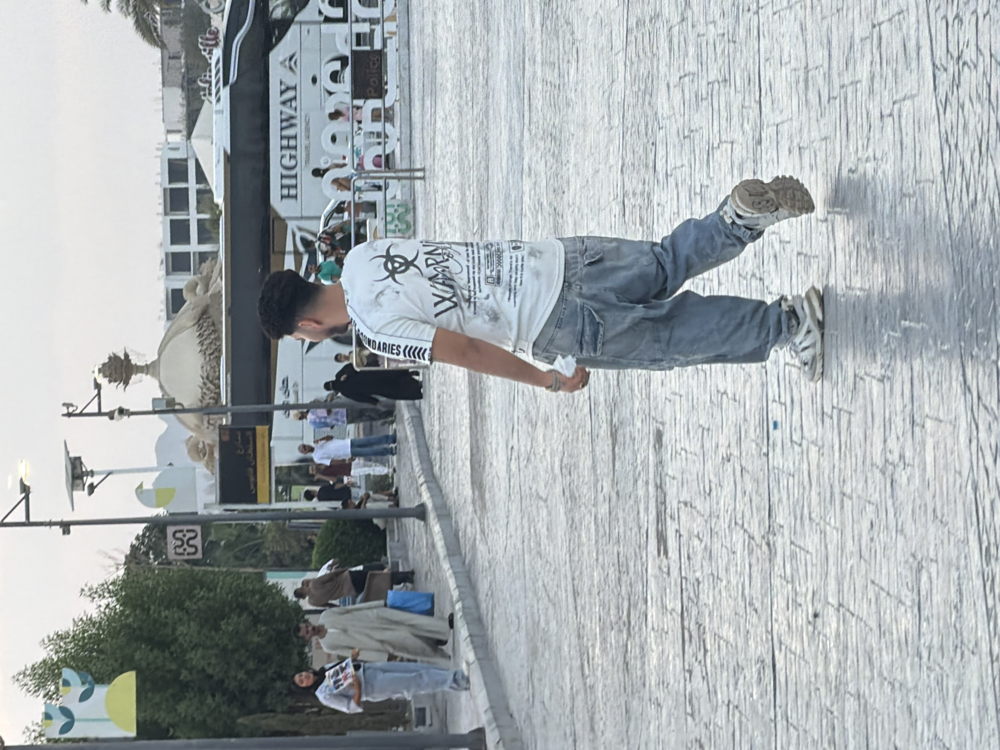 A young man with dark, curly hair walks away from the camera on a wide, paved pedestrian walkway in Sharm El Sheikh, Egypt. He is wearing a white t-shirt with a graphic design and text, and loose-fitting, light-wash denim jeans. His shoes are grey and white athletic sneakers. In the background, a large tour bus with "HIGHWAY TRAVEL AGENCY" written on its side is parked. Other people are visible in the background, some walking and others standing, with palm trees and buildings suggesting an urban environment. Streetlights with solar panels are present along the walkway. The overall lighting suggests it is either early morning or late afternoon.