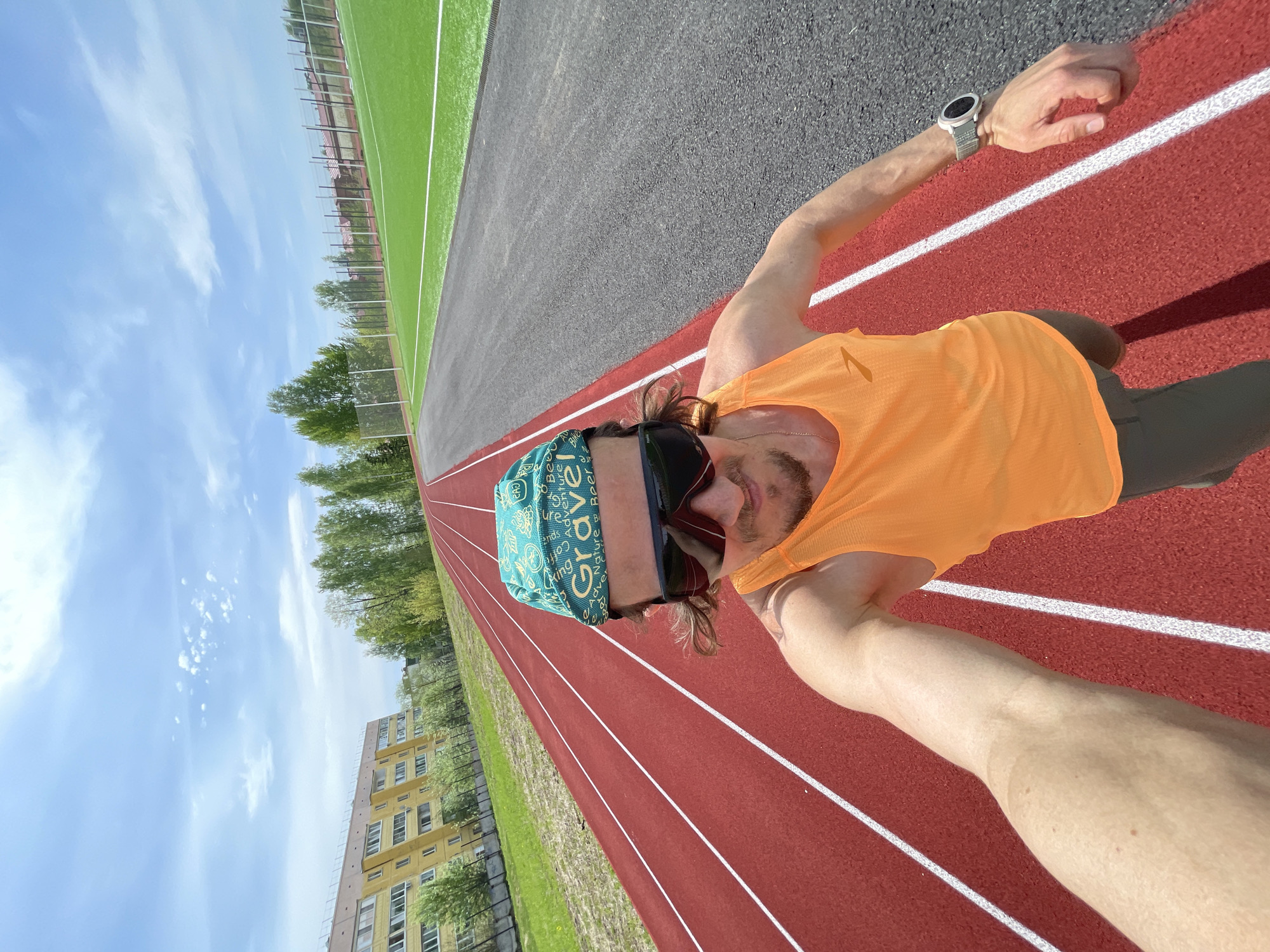 A selfie photograph features a light-skinned male wearing an orange sleeveless shirt, a teal bandana, and sunglasses on an outdoor running track. He is also wearing a watch on his left wrist. The running track, marked with white lines, is red with a gray area to the left. Beyond the track, there's a green field, trees, and an apartment building under a bright, slightly cloudy sky. The photograph appears to have been taken in Noviy, Russia, as stated in the instructions.