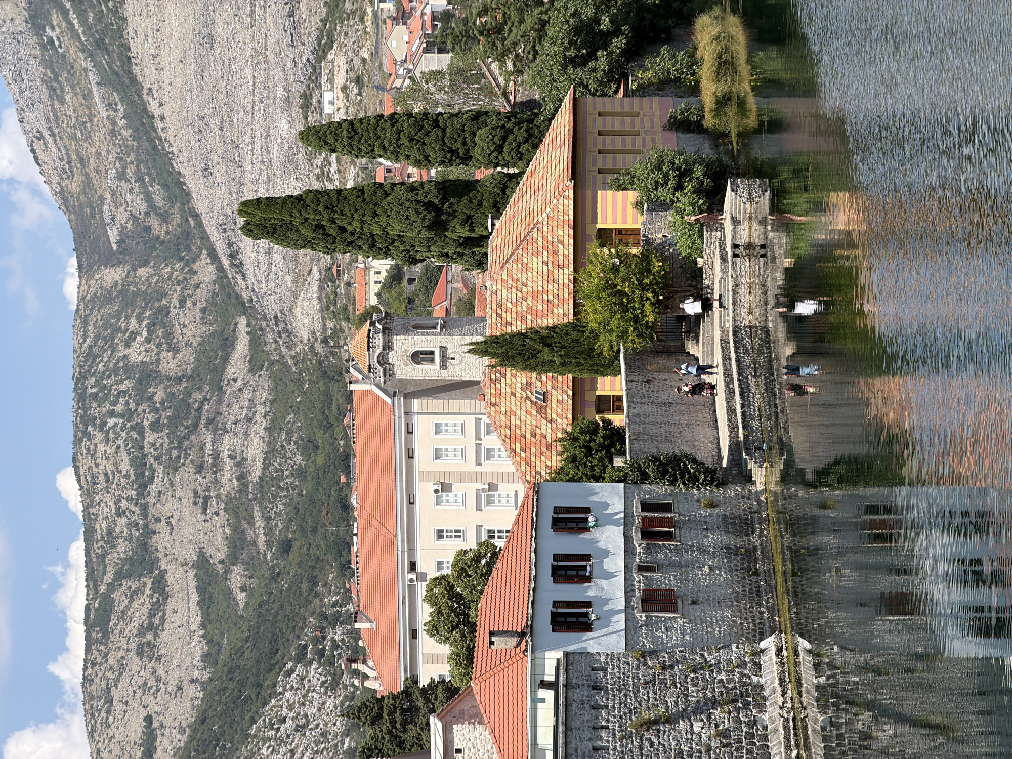 The image depicts a waterfront scene in Trebinje, Bosnia and Herzegovina, with buildings and a mountainous backdrop. In the foreground, a body of water reflects the surrounding architecture. On the left, a stone building with red shutters lines the water's edge. To its right, a smaller, yellow and pink building with a red-tiled roof is situated. Behind these, a larger, pale yellow building with a red-tiled roof is visible, and further back, a tower with a belfry stands out. The background is dominated by a large, rocky mountain, sparsely vegetated with trees and shrubs. Several people are visible walking along the waterfront. The sky is blue with scattered clouds.