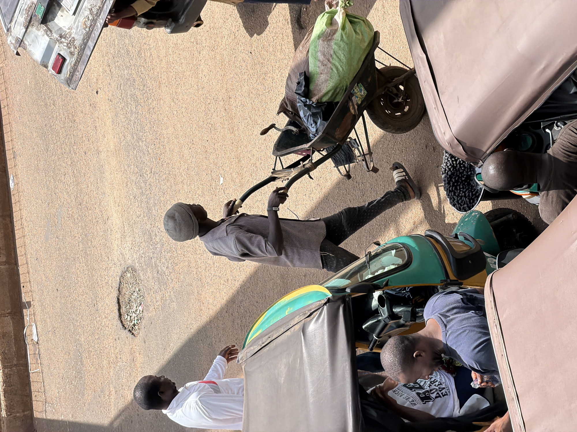 In Bukuru, Nigeria, a street scene unfolds with individuals and vehicles amidst a dusty, asphalted roadway. A man wearing a white shirt with a red stripe stands near the left edge of the frame, his gaze directed forward. To his right, another man in a grey, patterned shirt and dark trousers pushes a wheelbarrow loaded with two large sacks, one a green tarp-like material and the other a brown bag. The wheelbarrow has a visible metal frame and a single wheel. In the foreground, a portion of a green and yellow motorized rickshaw is visible, its canopy a dark brown. A man with short, dark hair, wearing a dark t-shirt, is seated in or near the rickshaw, holding an item in his right hand. Another individual, wearing a white t-shirt with text, is partially visible behind him. To the right of the wheelbarrow, a metal bin filled with dark, round objects, possibly olives or berries, sits on the ground. A portion of a rusty, weathered vehicle with taillights is visible in the upper right corner. The roadway shows signs of wear, including a noticeable pothole filled with debris. The overall impression is one of daily life and commerce on a Nigerian street.