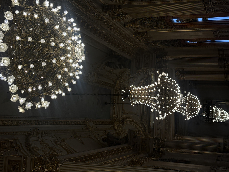 The image displays the opulent interior of the Salón Dorado (Golden Hall) at the Teatro Colón in Buenos Aires, Argentina. The scene is dominated by multiple illuminated chandeliers set against a dark, richly decorated backdrop. In the upper right quadrant, a large, ornate chandelier constructed of dark bronze or gilded metal features a dense array of spherical incandescent bulbs, some housed within white floral-shaped shades, along with a smaller crystal chandelier component at its core. Extending diagonally into the background are three additional multi-tiered crystal chandeliers, decreasing in size, each brightly illuminated and suspended by dark chains. The architectural environment includes dark walls adorned with extensive gilded moldings, tall fluted columns with gilded capitals, intricate friezes, and a decorated ceiling featuring faint ornamentation and possible murals. Arched window openings with dark frames and a subtle blue light are visible on the right wall, suggesting an exterior or adjacent space. No individuals or active subjects are present.