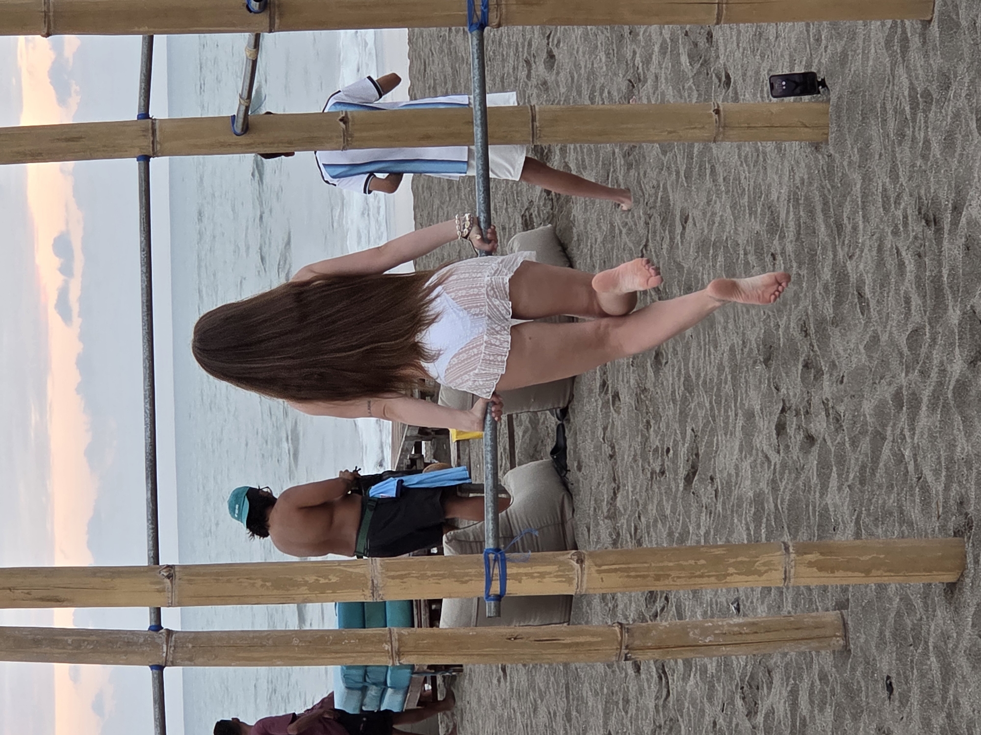A low-angle, full shot captures a beach scene in North Kuta, Indonesia, featuring several individuals and bamboo structures. In the foreground, a woman with long, dark brown hair is captured from behind, facing away from the camera and toward the ocean. She is wearing a white, lace-like bikini bottom and a sheer white cover-up skirt. Her left foot is raised, suggesting movement. To her left, partially obscured, is a man wearing a green cap and black shorts, shirtless. To the right of the woman, another individual is visible from the back, wearing a white t-shirt with blue vertical stripes and white shorts.
The environment consists of a sandy beach with the ocean visible in the background. The sky above is overcast with soft, muted colors, indicative of either dawn or dusk. The scene is framed by bamboo poles and a horizontal metal bar, which appears to be part of a structure. In the mid-ground, there are beanbag-like chairs and what appears to be stacked blue cushions on a wooden structure. A black smartphone on a tripod is positioned on the sand near the bamboo poles on the right side of the frame. The overall lighting suggests ambient daylight.