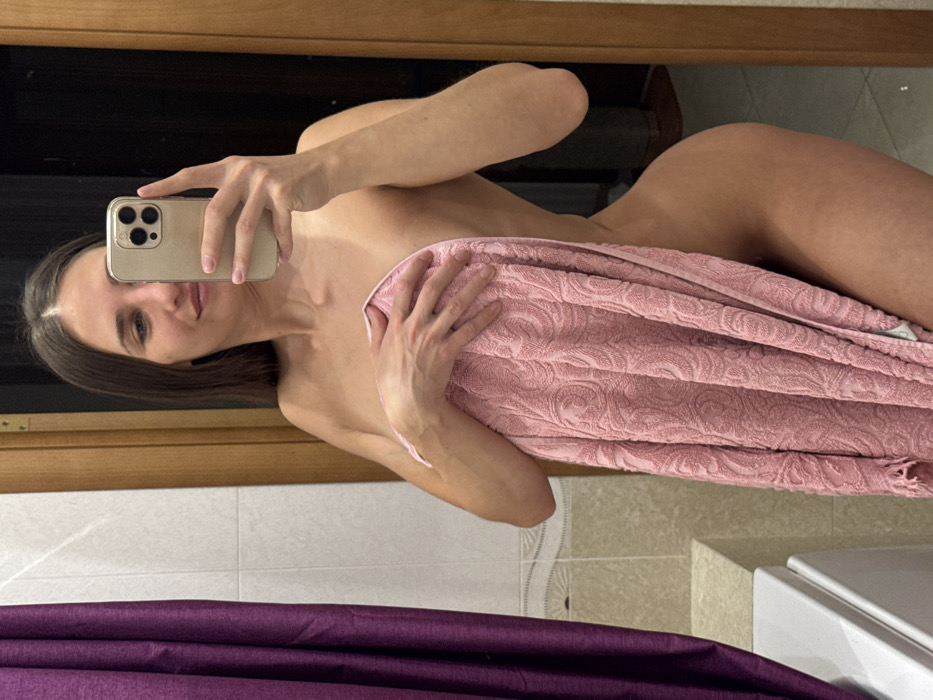 A woman in a bathroom is holding a pink towel and taking a mirror selfie with a modern iPhone. The bathroom has brown wooden trim, white tiles, and a purple curtain. The woman has long brown hair and is looking directly at the camera. The towel is covering most of her body.