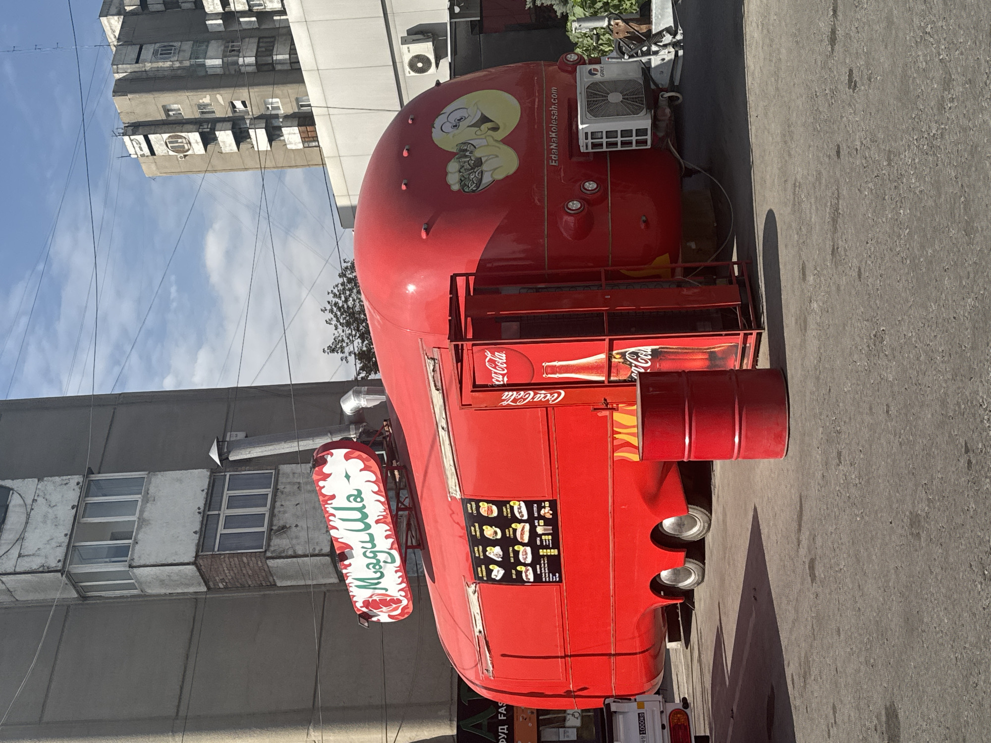In the city of Bishkek, Kyrgyzstan, a bright red food truck with the name "МагиШла" prominently displayed in stylized lettering sits parked on a paved surface. The truck's rounded body is decorated with flames on the rear section and a cartoonish yellow egg-like character holding a bowl of food on its side. A menu board featuring images and text in a foreign language is affixed to the side of the truck. Adjacent to the food truck, a red metal barrel stands upright. Behind the truck, a multi-story concrete building with balconies and windows is visible, and further in the background, another large apartment building looms against a blue sky with scattered clouds. The scene appears to be an outdoor setting, possibly a street or parking area.