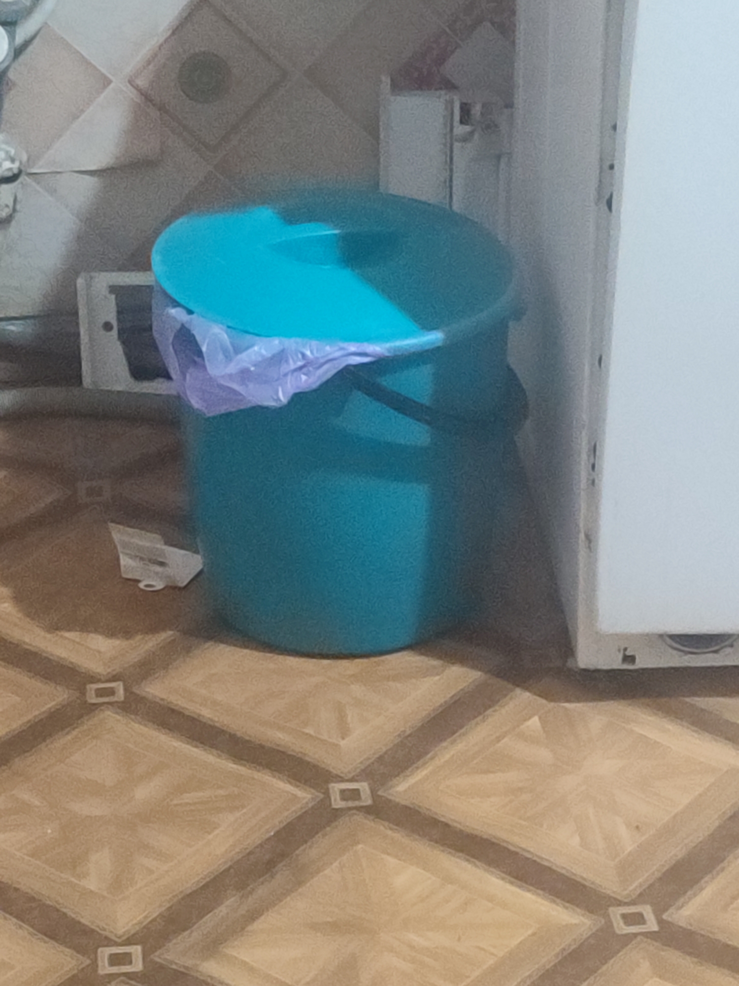 In Chernivtsi, Ukraine, a teal-colored trash can with a blue lid and a purple trash bag liner sits on a patterned tile floor. Behind the trash can, a white appliance, possibly a refrigerator, is visible. To the left of the trash can, a small white object is on the floor. The background shows a tiled wall.