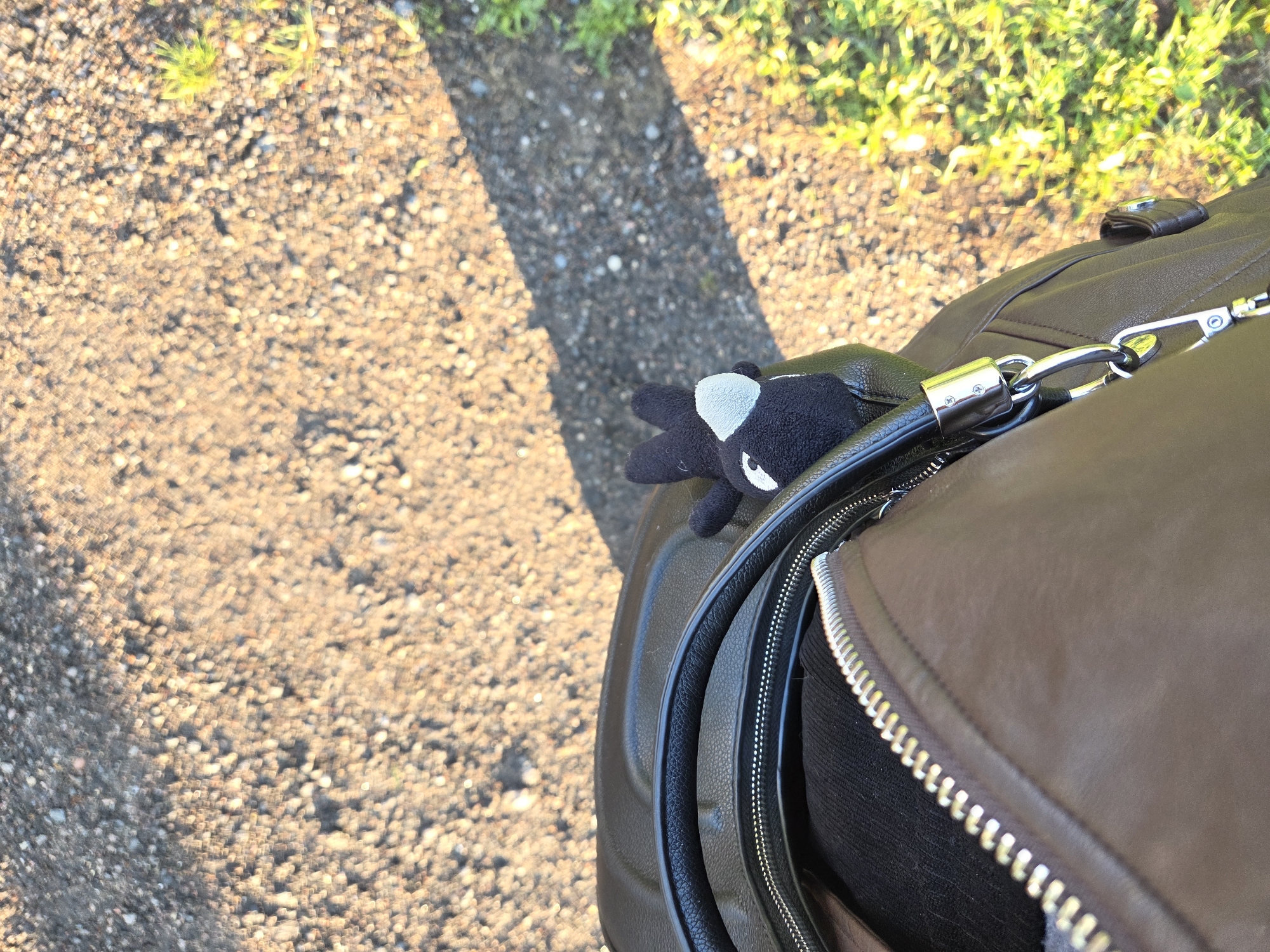 The image shows a portion of a brown leather bag with a black and white plush toy attached to its zipper. The bag is placed on a gravel or paved surface, with green foliage visible in the background. The setting appears to be outdoors.