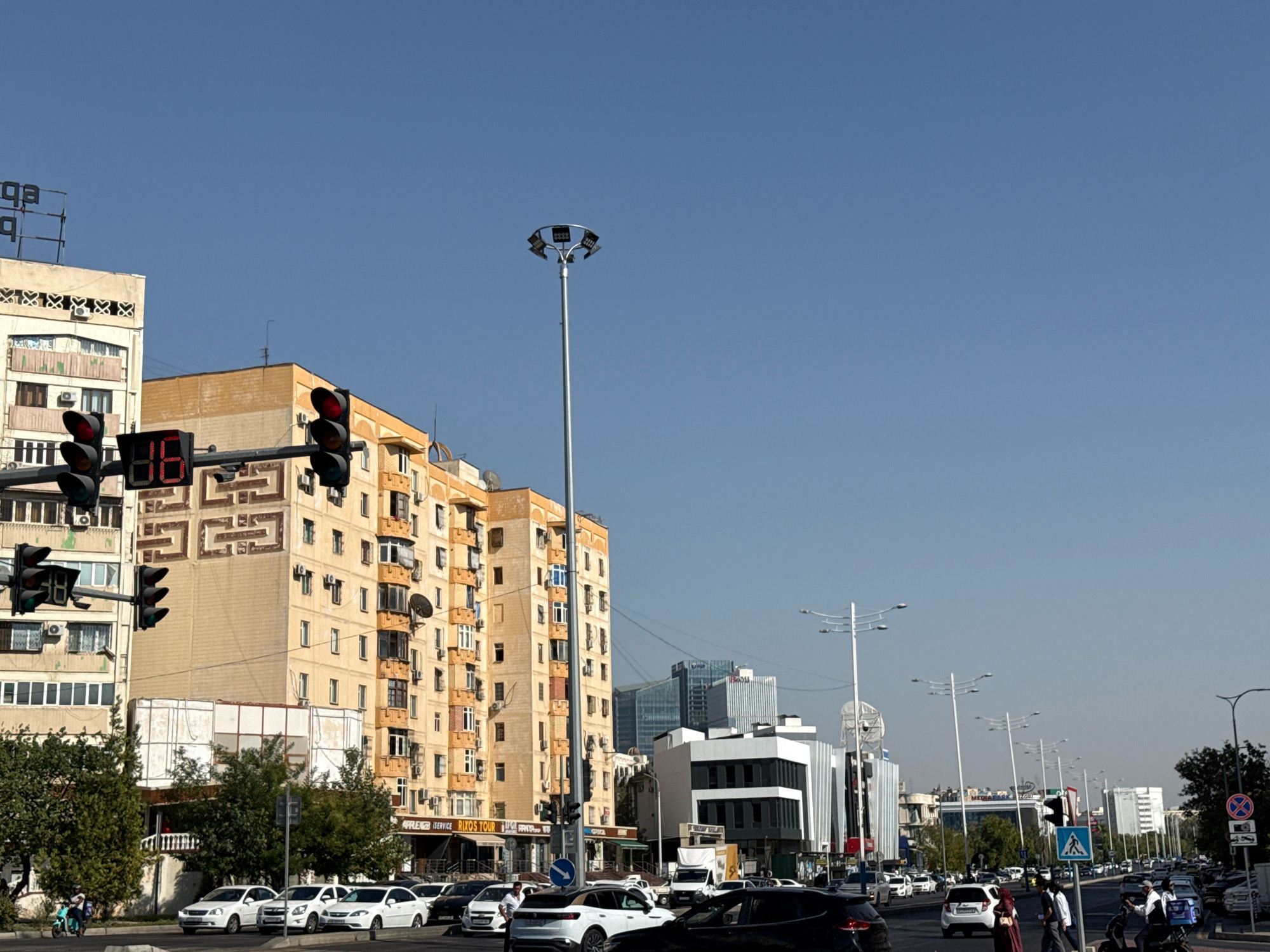The image captures a street scene in Tashkent, Uzbekistan, under a clear blue sky. In the foreground, a traffic light shows a red signal for cars. To the left, a multi-story building with a beige facade and white window frames stands prominently. Adjacent to it, another building, also beige, features ornate decorative elements above its windows. Further down the street, a traffic light pole with multiple lights is visible.
The middle ground reveals a dense urban environment with several buildings. A tall, yellow-toned building dominates the right side, its facade adorned with numerous windows and balconies. Below this building, shop signs, including "APPLE" and "RIXOS TOUR," are discernible. In the background, modern glass-fronted buildings rise, indicating a mix of old and new architecture.
Vehicles, including cars and what appears to be a scooter or motorcycle, are present on the road. Pedestrians are also seen, some crossing the street. The overall scene suggests a bustling urban thoroughfare with active traffic and commercial activity. The presence of numerous trees lining the sidewalk and interspersed among buildings adds a touch of greenery to the cityscape. The clear sky and bright sunlight indicate a pleasant day.