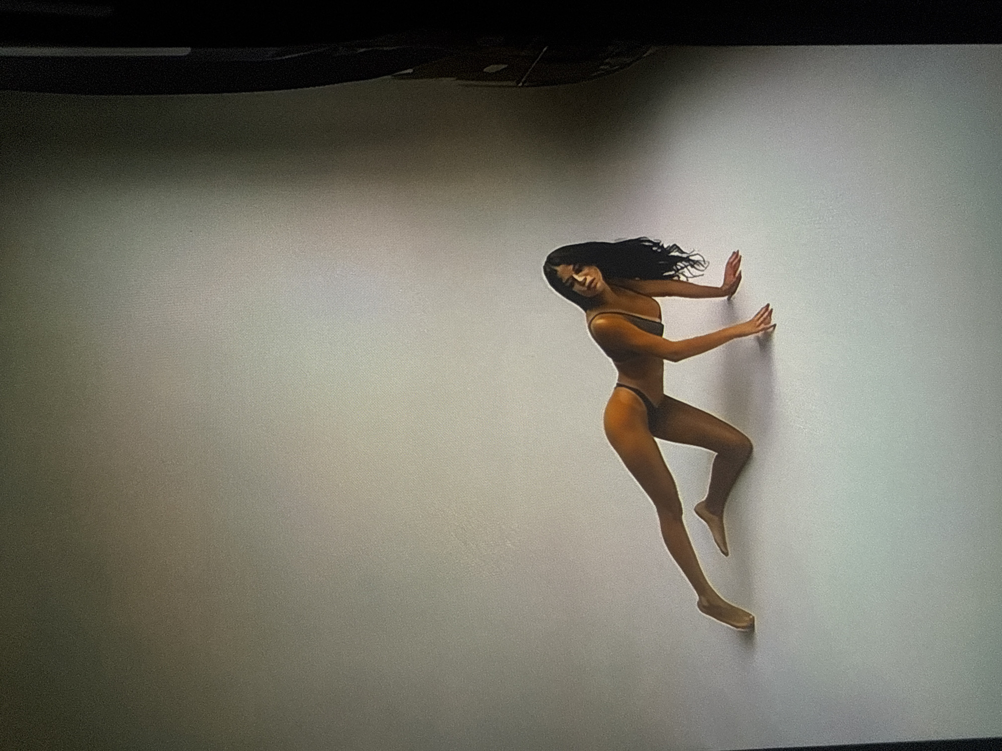 The image depicts a studio photograph of a single female subject. The woman has long, dark, wavy hair and a warm skin tone, and is wearing a dark two-piece garment, consistent with a bikini or lingerie. She is positioned in a low, dynamic pose, with her left knee bent and resting on the light-colored floor, and her right leg extended backward and slightly upward. Her hands are placed flat on the floor in front of her, providing support. Her head is tilted towards her right, and her gaze is directed away from the viewer. The background is a plain, seamless, light grey or off-white surface, characteristic of a professional photographic studio setting, implying controlled lighting. A dark, vertical element, possibly studio equipment or a backdrop edge, is visible along the far right side of the frame. The overall image exhibits a fine grain or digital noise. The described scene is located in the city of Varna, Bulgaria.