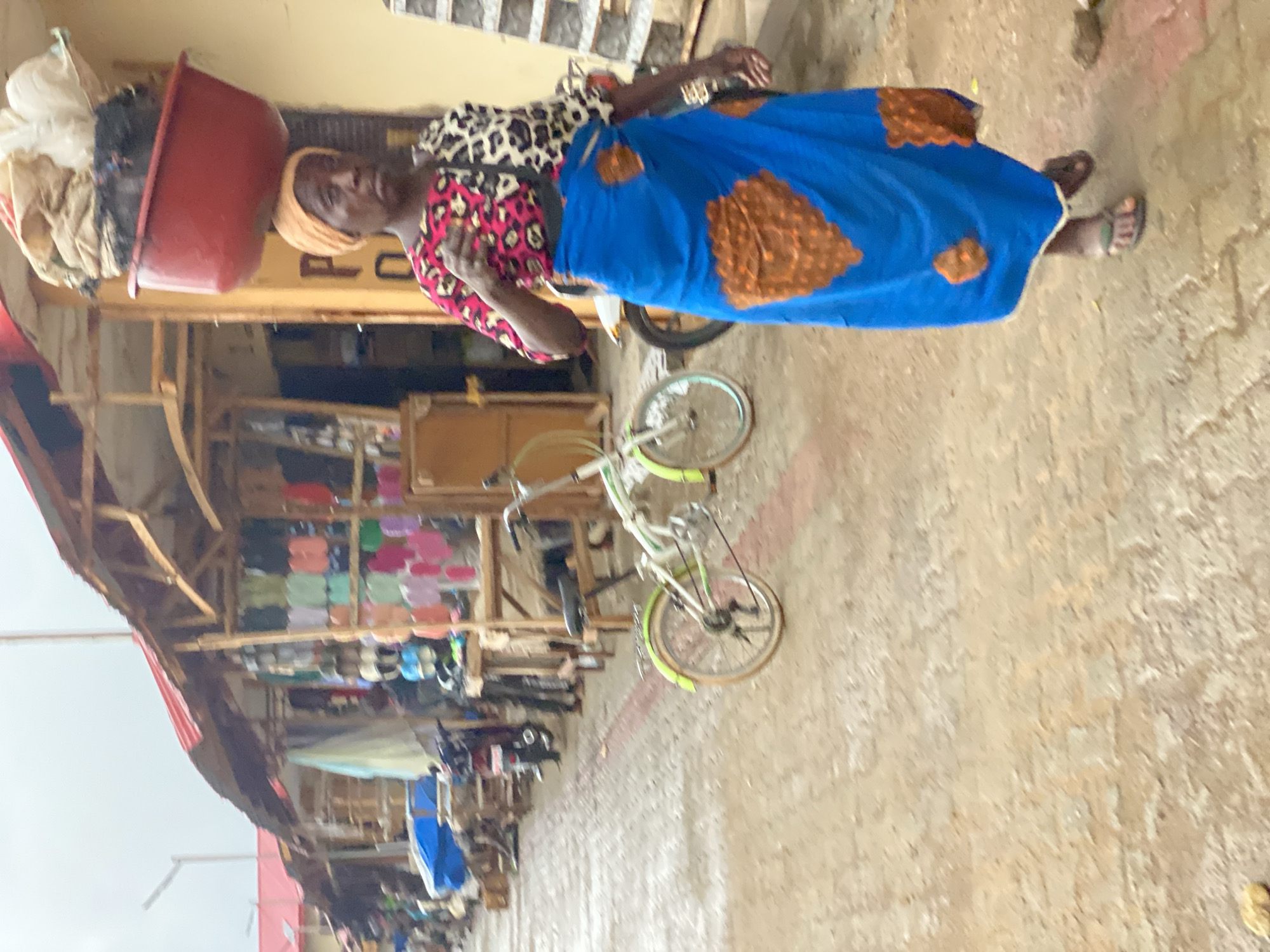 A woman stands in a market in Kafanchan B, Nigeria, with a red bowl balanced on her head filled with various items. She wears a patterned blouse and a blue skirt with brown designs. A bicycle rests against a wooden structure displaying assorted merchandise. The ground appears wet.