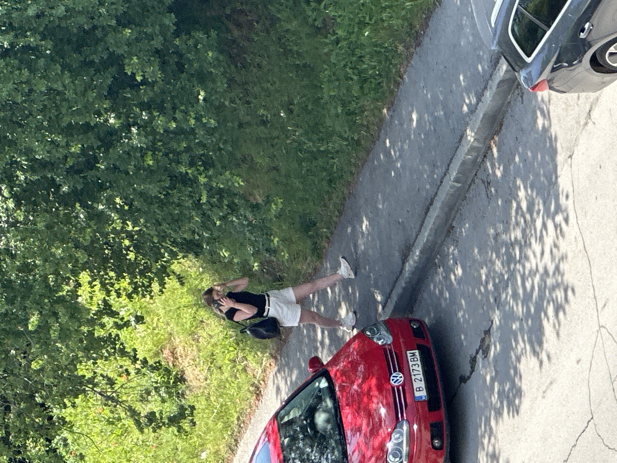 A high-angle shot depicts a woman standing on a sidewalk, apparently talking on her phone. She is wearing a black shirt, white shorts, white shoes, and has a black bag over her shoulder. She stands between a red Volkswagen car (license plate B 2173 BM) and a grass/tree area. A gray car is parked on the sidewalk next to the woman. The location appears to be along a street lined with trees, and the scene includes dappled sunlight. Given the license plate format (B followed by numbers and BM), the setting is likely in Romania, not Varna, Bulgaria.