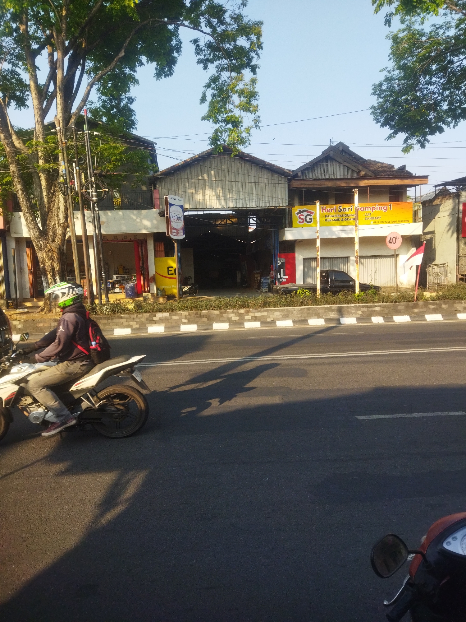 In Sukoharjo, Indonesia, a street scene unfolds under a clear sky, bathed in the warm light of what appears to be late afternoon or early morning. A person on a white motorcycle, wearing a helmet with green and white accents and a dark jacket over a backpack with red straps, rides along the asphalt road. The motorcycle has a visible engine and wheel.
Adjacent to the road is a sidewalk with a white and gray curb. Behind the sidewalk are several buildings. One building features a large yellow banner with red lettering that reads "Heri Sari Gamping 1" and "SG BAHA BANGUNAN - KERAMIK & GRANIT - CAT - SANITARY". Underneath this banner, a black car is parked in front of a building with closed roll-up doors. Another building, to the left, has a red and yellow facade with signs and merchandise visible inside. To the right of the yellow banner, a circular sign with a red border displays "40 km". A flagpole with a red and white Indonesian flag stands near this sign. Tall trees with green foliage and thick branches are prominent in the background, casting shadows on the road. Utility poles and wires are visible near the buildings and trees. A portion of another motorcycle, orange in color, is visible in the lower right corner of the frame.