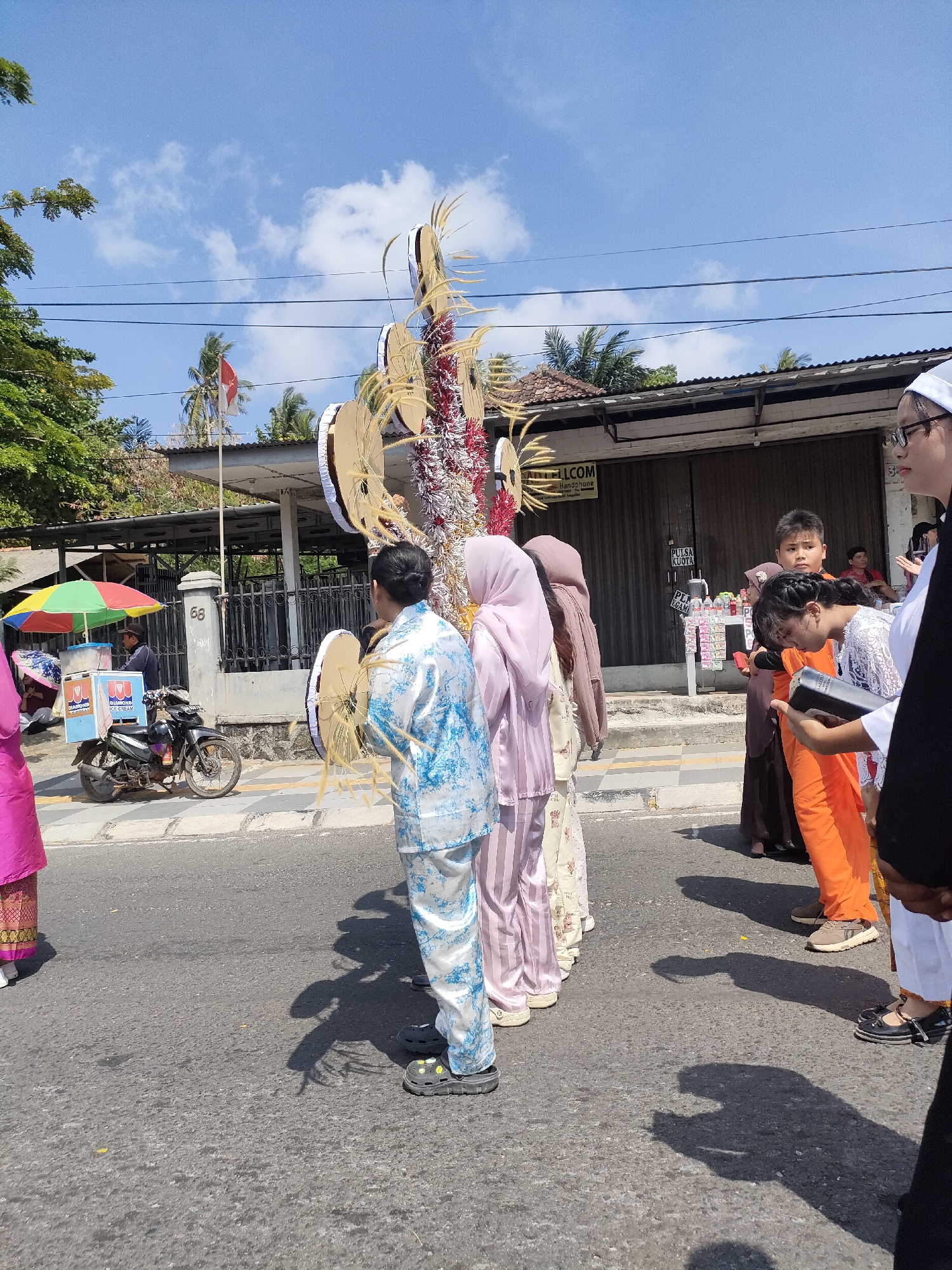 The image depicts a street scene in Sungailiat, Indonesia, under a clear blue sky with scattered clouds. A group of people, some wearing traditional Islamic attire, are gathered on the road. In the foreground, a person in a white and blue patterned outfit stands facing away from the camera, holding a large, ornate, gold-colored decorative item adorned with red and white tinsel and wheat-like plumes. To their left, another person in a pink outfit is partially visible.
Behind the central figure, several other individuals are seen, including a person in a light purple hijab and a patterned dress, another in a brown outfit, and a young boy in a brown tunic. To the right, a young girl wearing an orange garment and a white lacy top holds a dark book. Further to the right, a person in a white religious head covering and glasses looks towards the left.
In the background, a building with large wooden doors and a sign that appears to read "ADICELL.COM" is visible. In front of the building, there are shelves displaying various items. To the left of the building, an ice cream cart with colorful umbrellas is set up next to a motorcycle. A fence separates the street from the building. The overall impression is of a street procession or gathering.