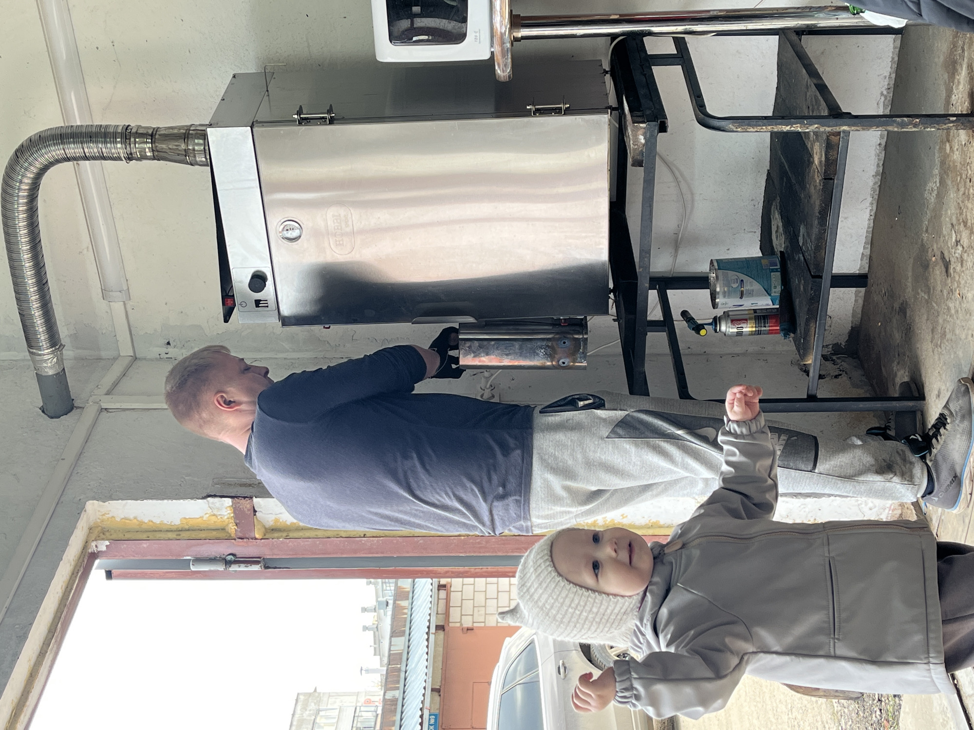 A man in a navy blue long-sleeved shirt and gray sweatpants is working on a stainless steel oven or smoker in an indoor setting. The oven has a temperature gauge and controls on the front and a flexible metal exhaust pipe connected to the top. The man is wearing black gloves. Beside him, a baby in a gray outfit and a white knit hat is standing with its hand raised. The background includes a metal frame, a can of paint or solvent, and a spray can. Outside a doorway or window, buildings are visible, including an orange building with signage in Russian. Based on the signage in Russian, the location is in the city of Chaikovsky, Russia.