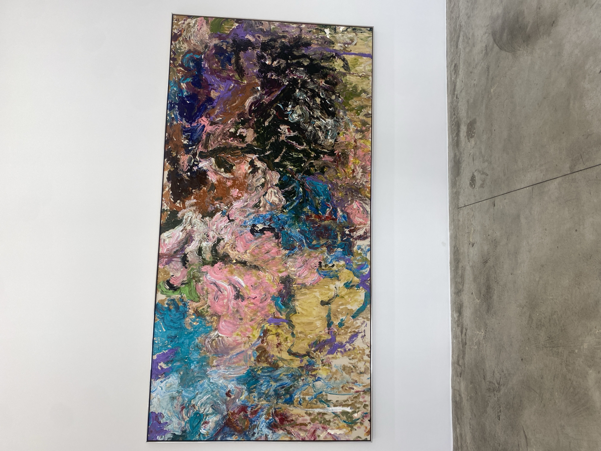 The image shows an abstract painting hanging on a white wall, with a concrete wall visible to the right. The painting is predominantly composed of strokes of various colors including pink, blue, purple, brown, green, black, and gold. The painting is framed with a thin metallic frame. No people are visible. The location cannot be determined based on the image.
