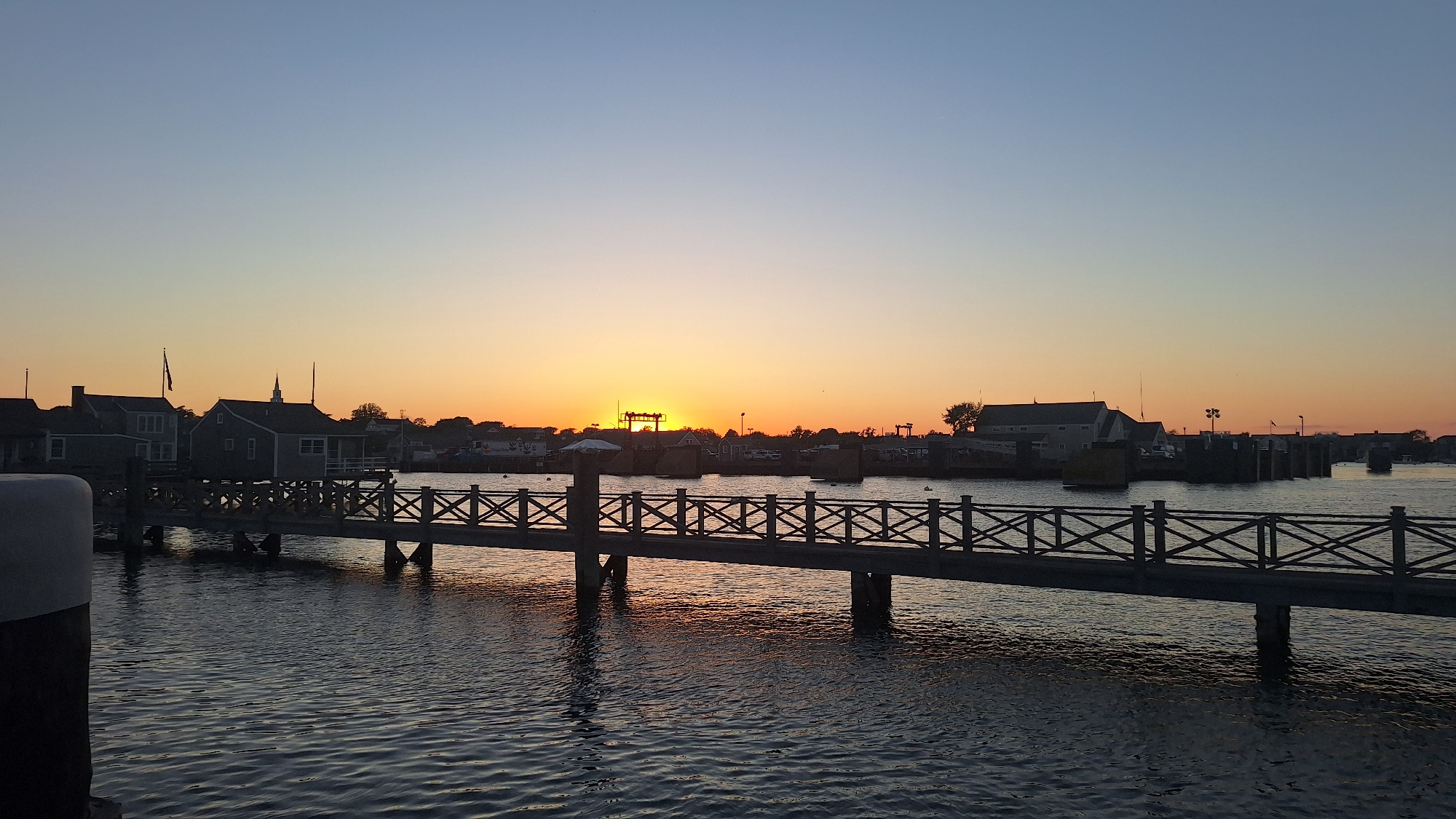 A wide, horizontal, eye-level shot shows a serene sunset over a harbor in Nantucket, Massachusetts, United States of America. The sky transitions from a clear, light blue at the top to a soft orange hue near the horizon. The sun, a bright orb, is setting behind a low-lying treeline and buildings on the far shore, casting a warm glow across the scene.
In the foreground, a dark, wooden pier with an intricate crisscross railing extends from the left side of the frame towards the center. The pier is supported by thick, dark pylons that cast reflections on the rippling water. The water itself is a dark, deep blue, reflecting the colors of the sky and the pier.
Along the opposite shore, silhouetted buildings, some with peaked roofs and a steeple, are visible. The architecture suggests a historic New England coastal town. A few boats are moored in the harbor, their shapes indistinct in the fading light. The overall atmosphere is peaceful and tranquil, characteristic of a quiet evening by the sea.