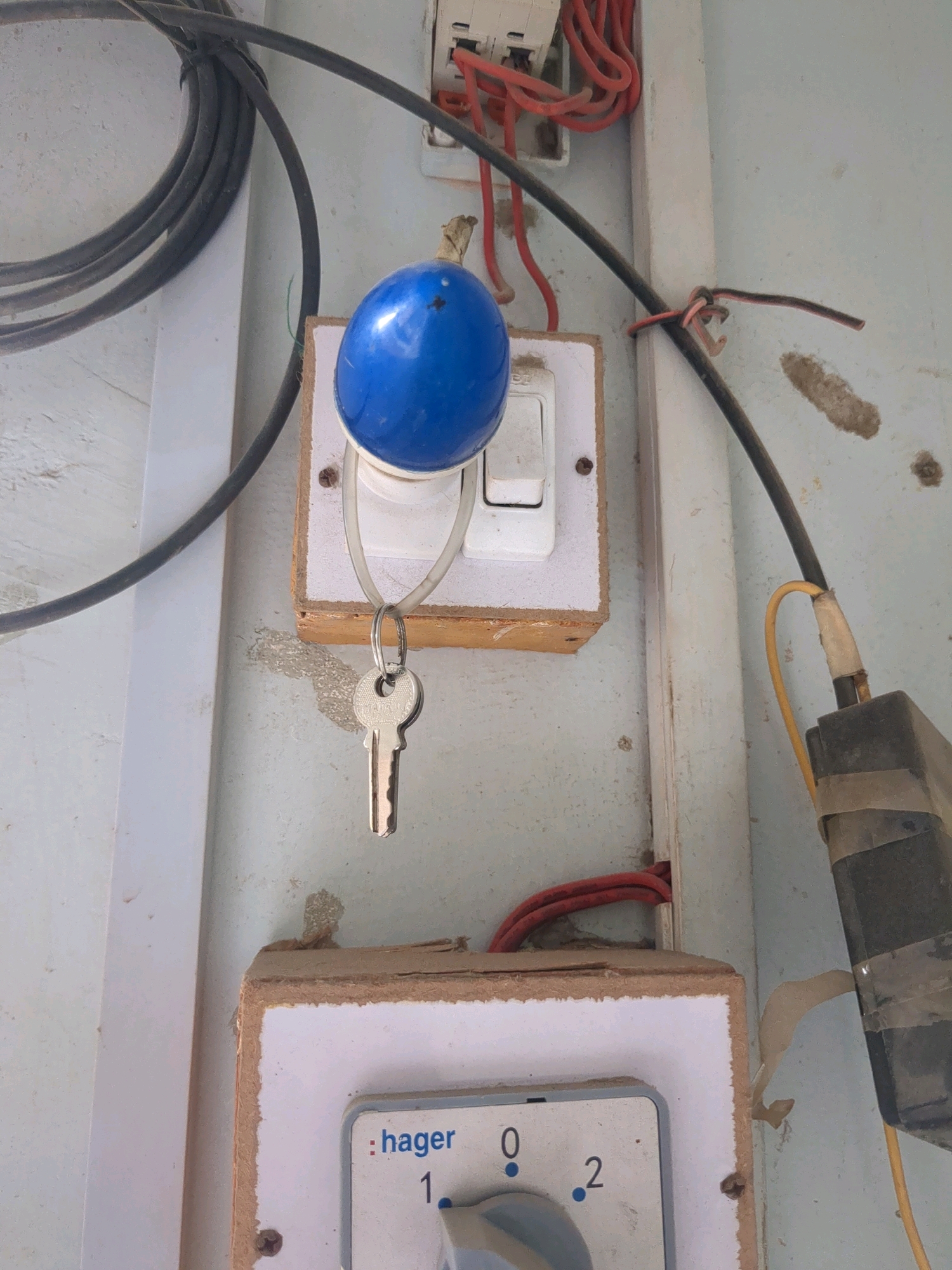 An outdoor close-up shot shows electrical components and wiring against a pale, textured wall. In the upper half, a white electrical switch with a blue knob and a key hanging from it is mounted on a light brown wooden panel. Red wires are visible connected to a white electrical box above the switch. A thick black cable coils in the upper left corner. To the right of the switch, a black electrical component is taped to the wall with yellow and grey tape, connected by a yellow wire. In the lower half, a second, larger electrical switch with the "hager" logo and numerical markings "0", "1", and "2" is also mounted on a light brown wooden panel. Red wires are visible entering this panel from the right. The wall shows some discoloration and damage. The scene appears to be in a utility area or an outdoor electrical junction box.